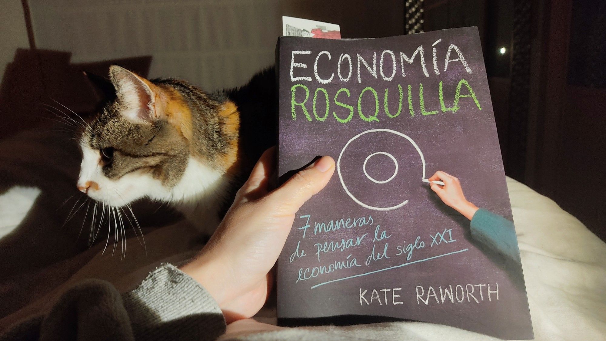 Mi mano izquierda sosteniendo el libro "Economía Rosquilla" de Kate Raeorth. Detrás del libro asoma Neka, mi gata.