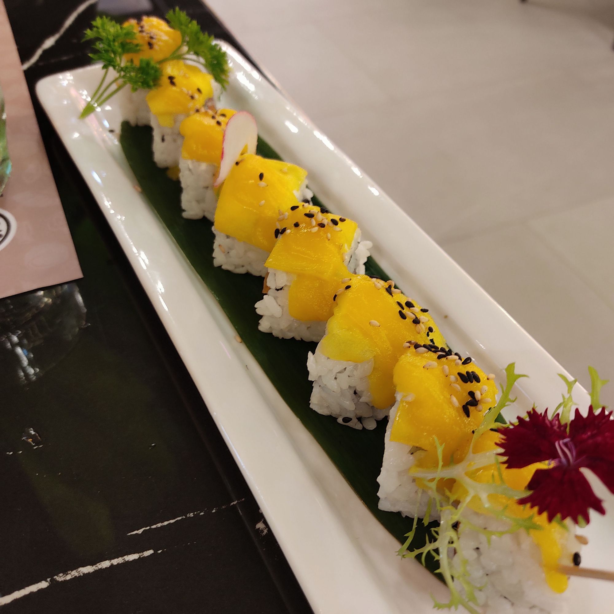 Plato de sushi de salmón con mango presentado todo cortado en ocho piezas con en mango por encima y unas flores pequeñas como decoración del plato.