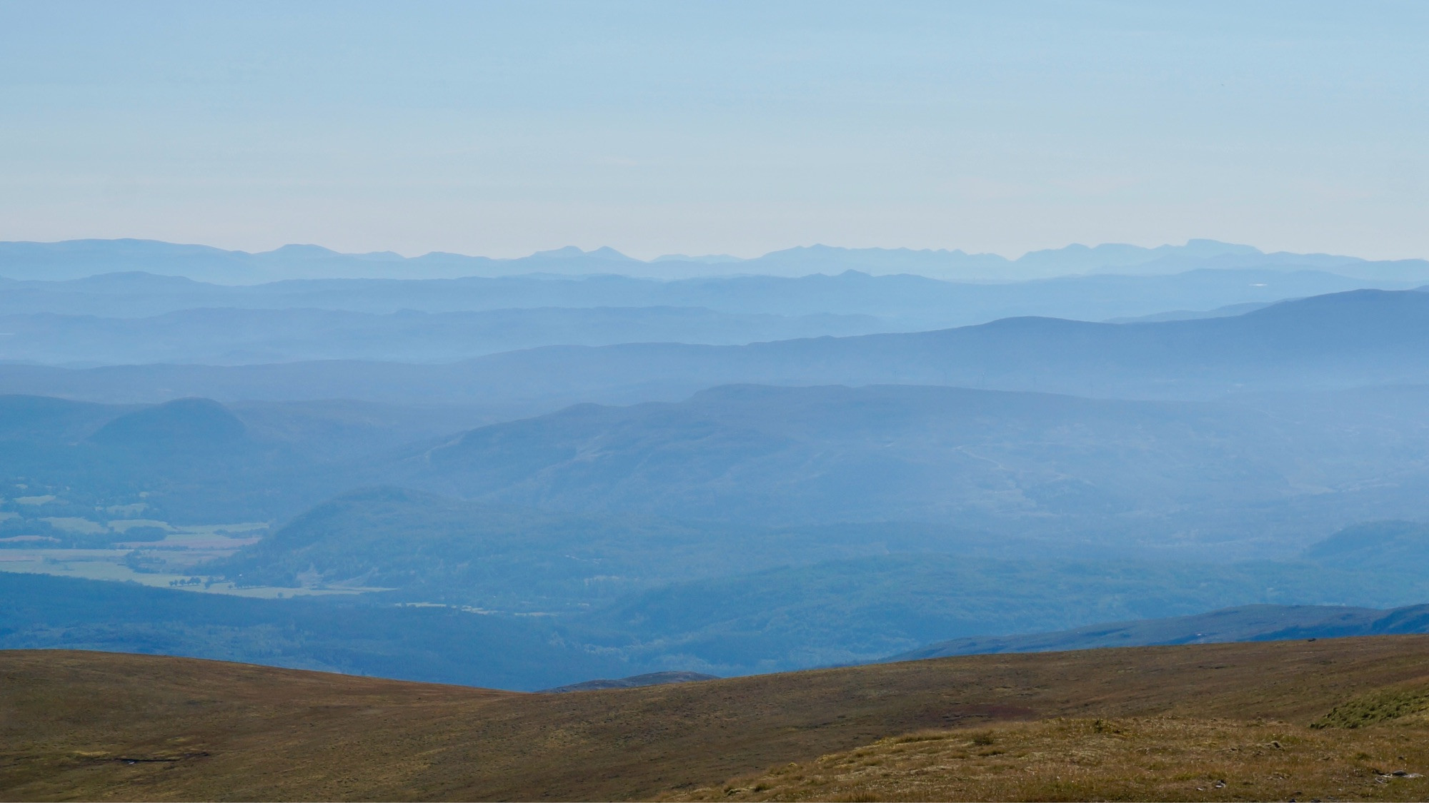 Ridges recede into the distant horizon, from closer deep dark blue to light grey before merging into the afternoon light sky