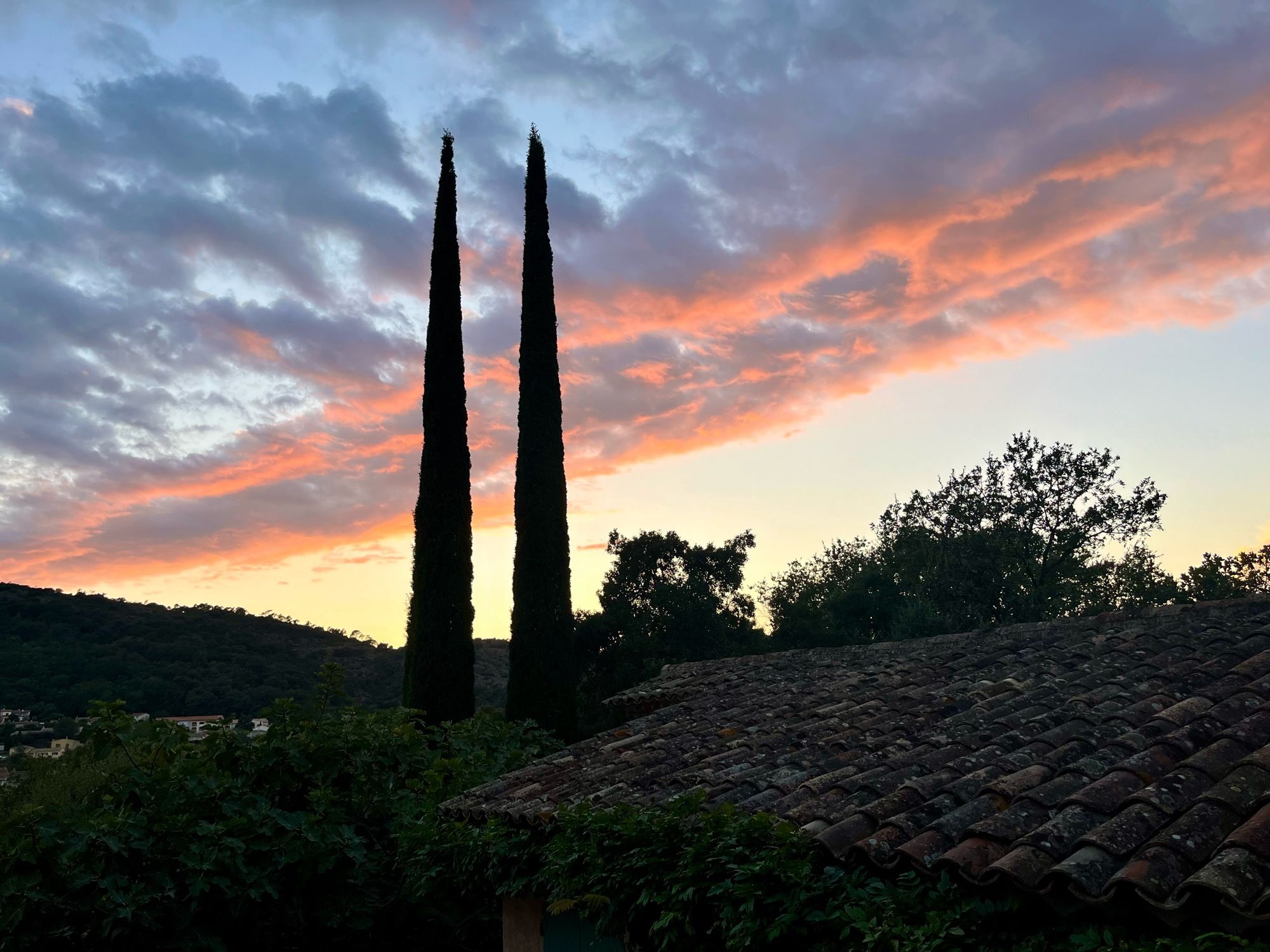 Avondlucht in de Provence. Van midden links tot rechtsboven een band wolken, blauw met veel oranje. Rechts daarnaast nog wat lichtblauwe lucht. Links van het midden doorsnijden twee hoge cipressen de wolkenbank. Rechts vooraan, in het donker, een Provençaals pannendak.