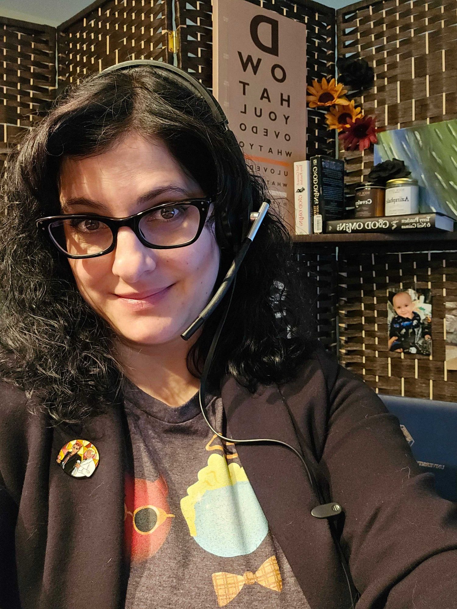 Picture of me in my Good Omens shirt, lapel pin, books behind me, and Aziraphale and Crowley candles