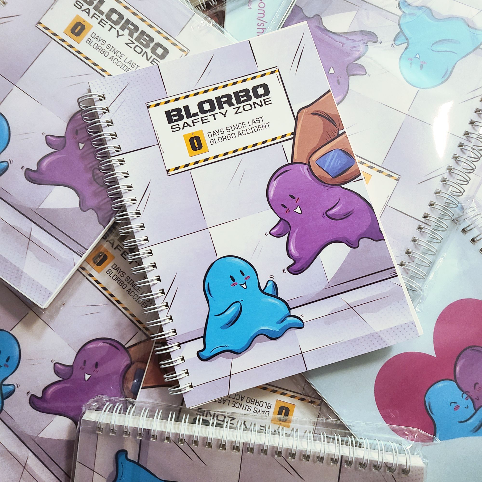 a spiral notebook with art on the cover of a blue blobby figure smiling and reaching out to a purple blobby figure, who is being set down by a giant light brown hand. a sign on the wall behind them reads "blorbo safety zone | 0 days since last blorbo accident"