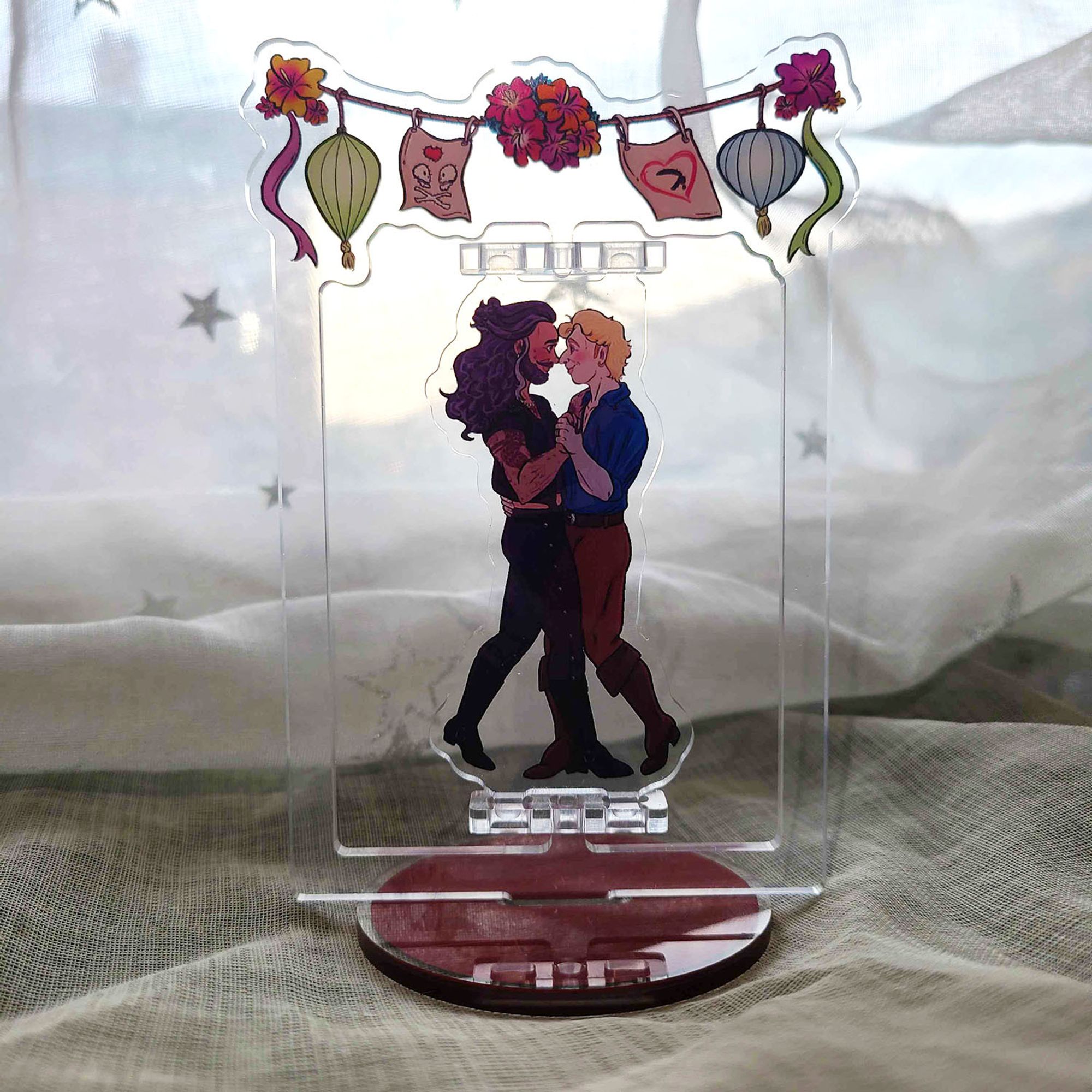 Acrylic standee of prev art: Ed and Stede are dancing together, looking into each other's eyes and smiling. Above their heads are flowers, paper lanterns, and handmade pirate flags. They are wearing their outfits from the Calypso's birthday party, but they're also both wearing gold rings on their left ring fingers, and Stede's ear is pierced.