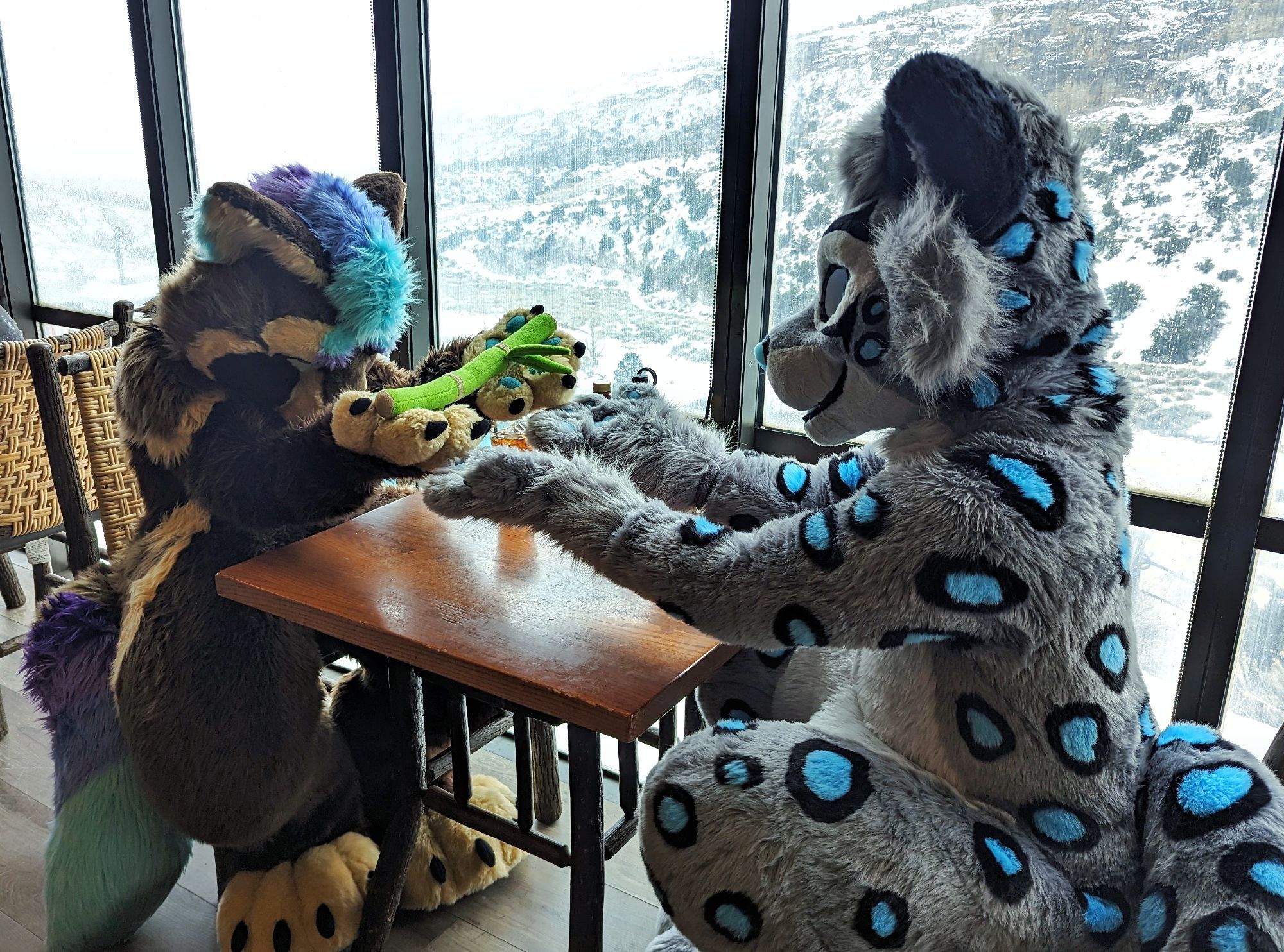 Satchel in fursuit, holding out his bamboo as a peace offering with both paws to Mihlo the snow leopard sitting across a table. Outside is a snowy hillside dotted with trees.