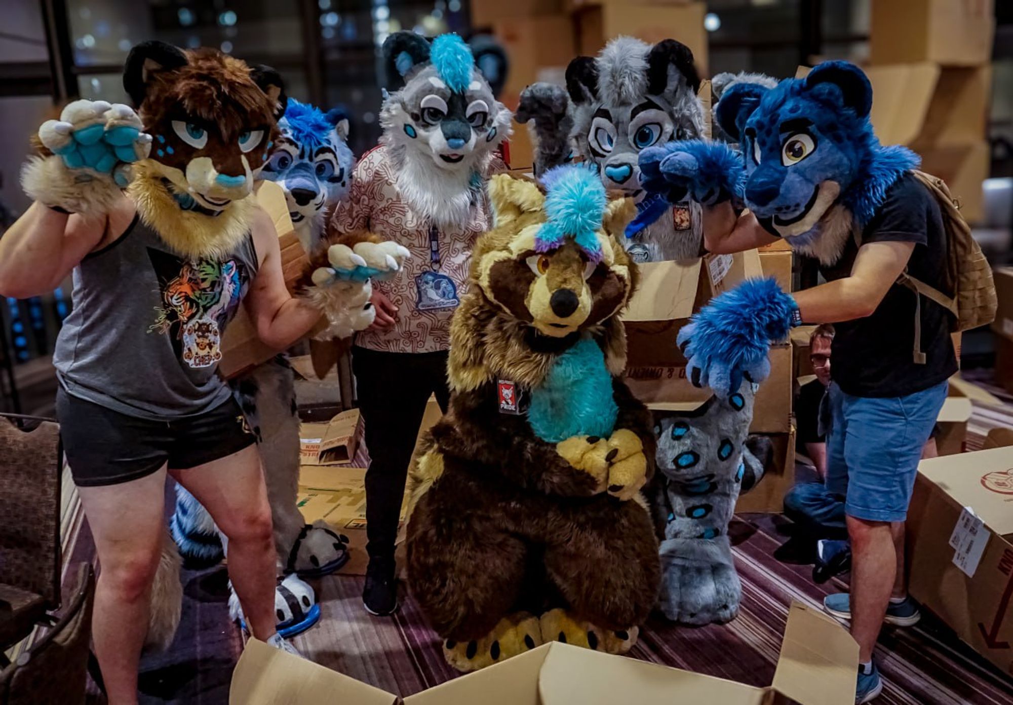 Satchel in fursuit, surrounded by snow leopard fursuiters amongst a group of cardboard boxes. He has his typical, mildly frightened expression on his face.