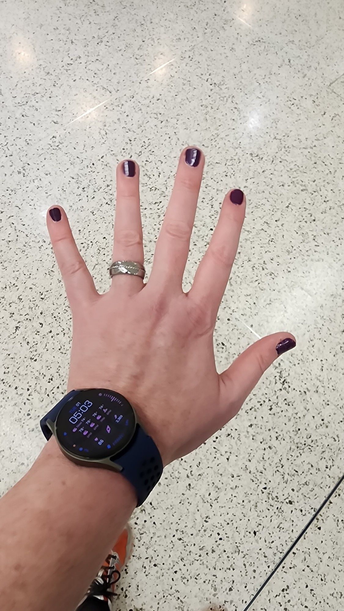 Left hand palm down fingers extended, purple nails, wedding band, smartwatch, orange shoe peeking in background