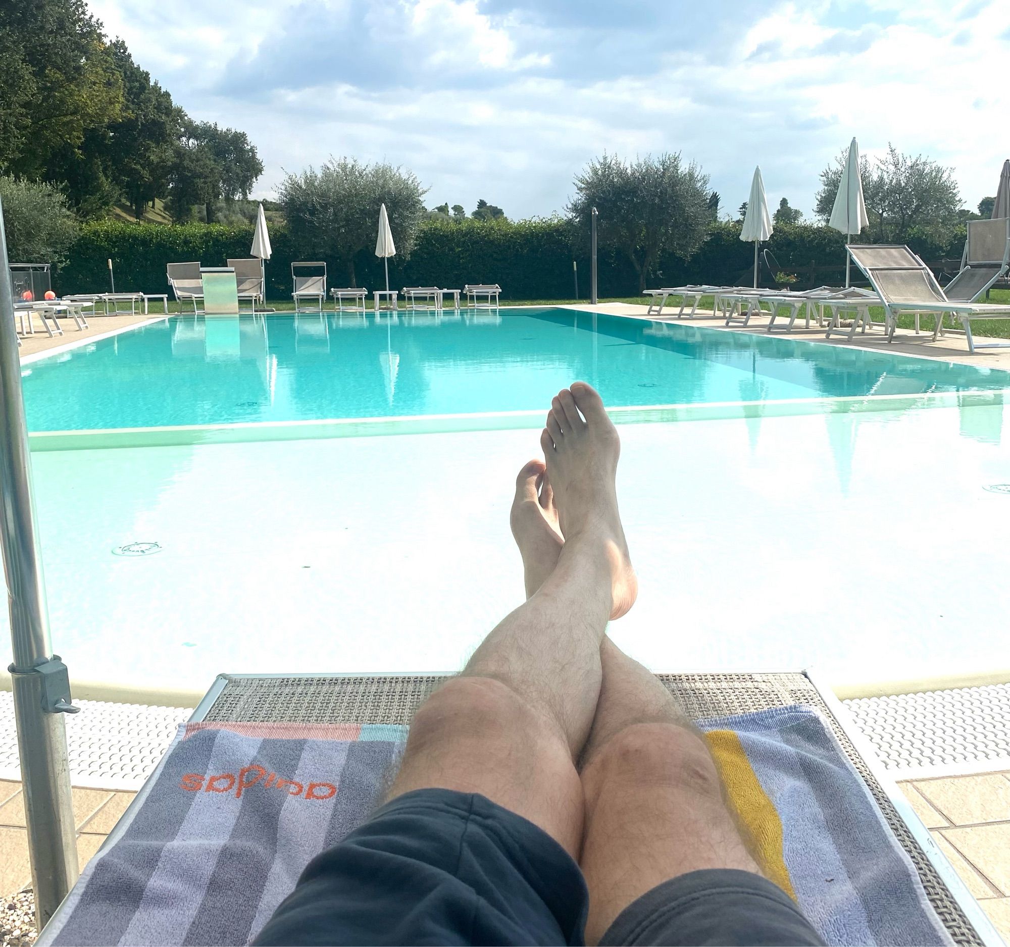 Auf der Sonnenliege, ein Blick über meine beschatteten Beine hinweg, auf einen Swimmingpool in der Sonne.