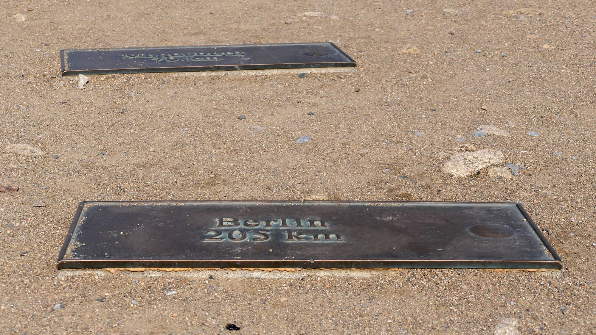 Das Bild zeigt zwei rechteckige Metallplaketten im Boden. Auf der vorderen Plakette steht "Berlin 205 km", was die Entfernung nach Berlin angibt. Der Boden besteht aus Sand oder Kies, und die Plaketten sind leicht eingesenkt.