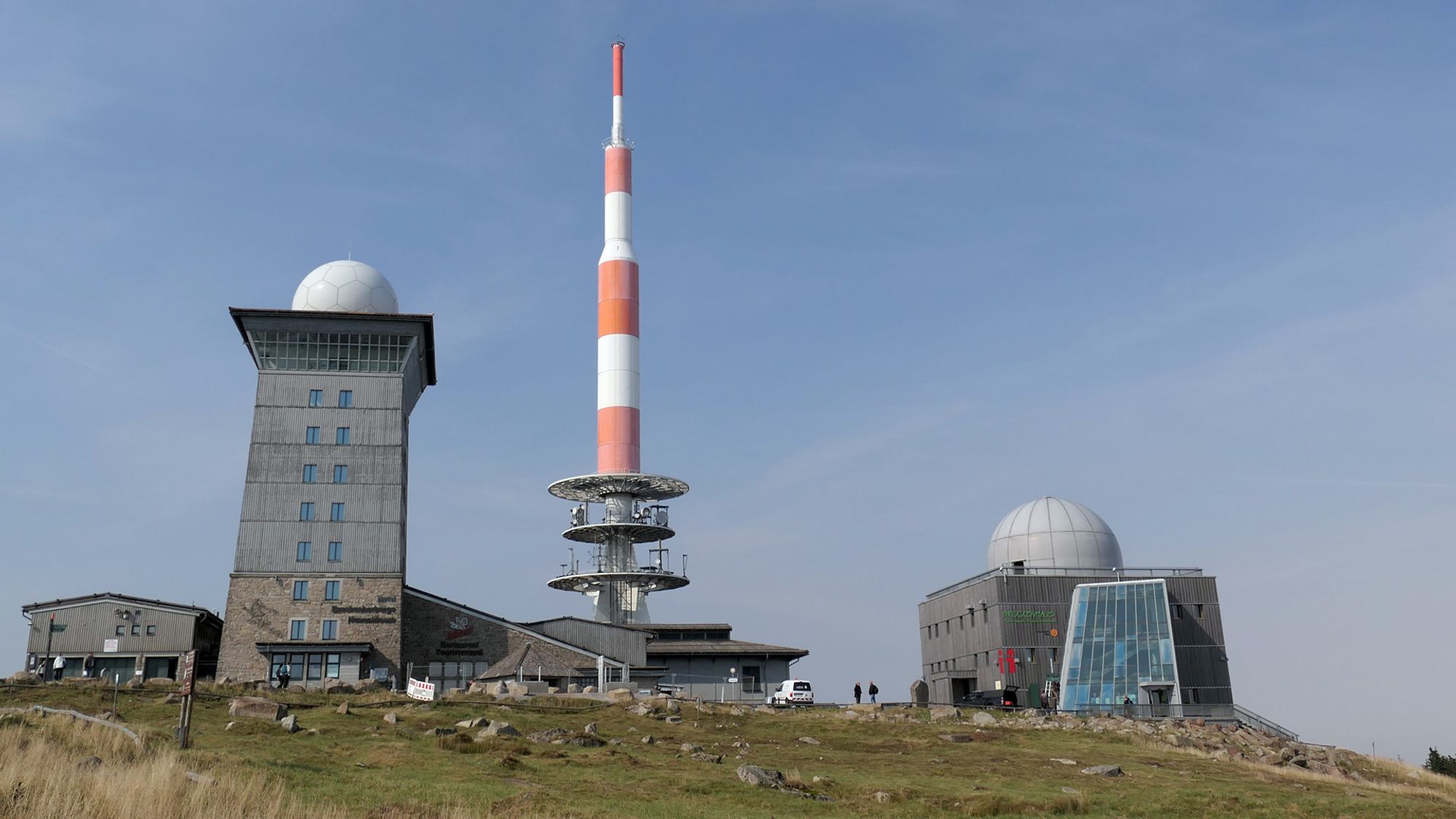 Das Bild zeigt den Gipfel des Brockens mit den markanten Gebäuden des Brockenobservatoriums. Im Zentrum steht ein hoher rot-weiß gestreifter Sendemast. Rechts und links davon sind moderne Gebäude mit großen Kuppeln zu sehen, die zur Wetter- und Forschungsstation gehören. Der Himmel ist klar und blau.