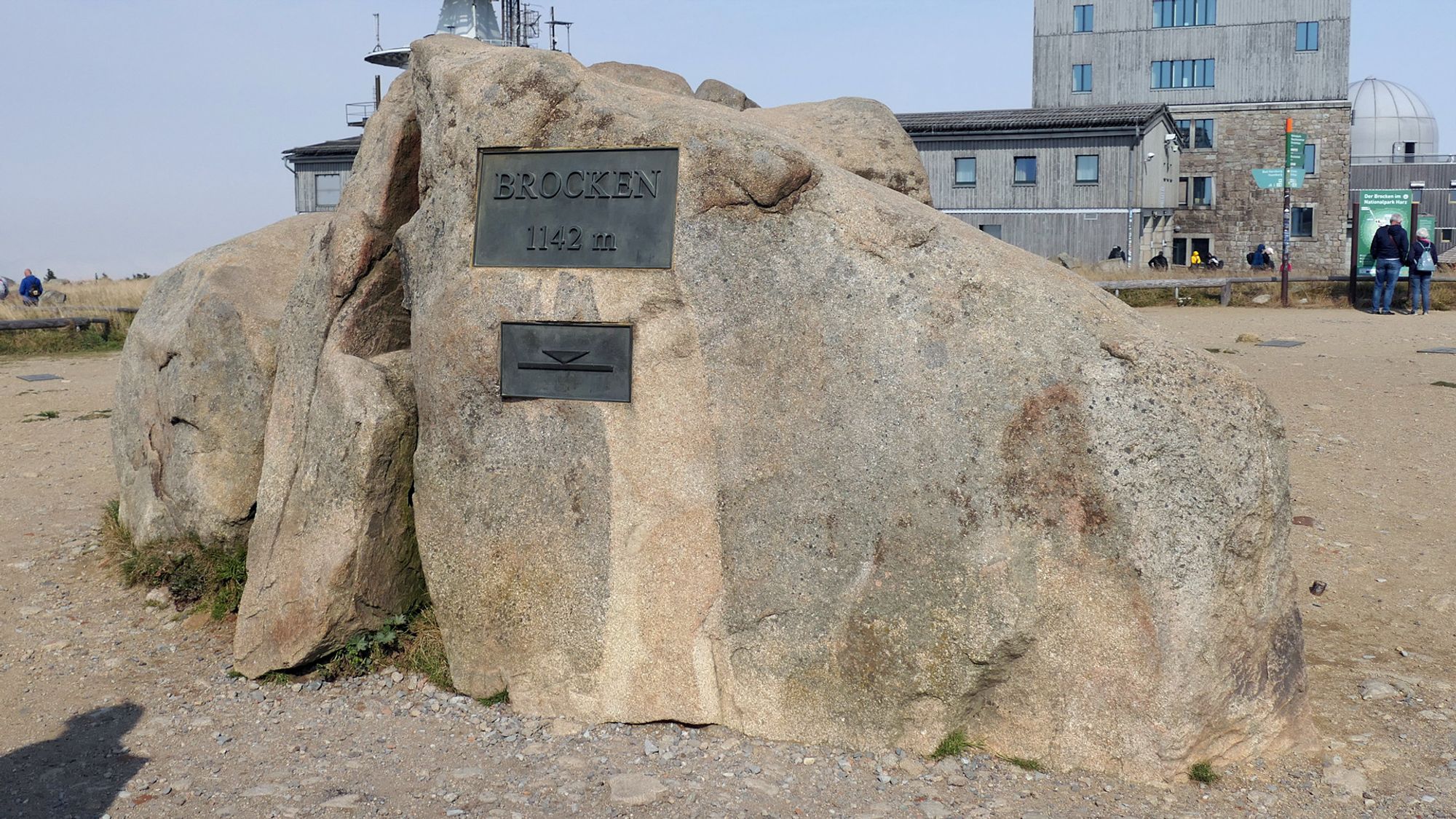 Das Bild zeigt einen großen Felsen auf dem Gipfel des Brockens, auf dem eine Metallplakette mit der Aufschrift "Brocken 1142 m" angebracht ist. Im Hintergrund sind Gebäude des Brockenobservatoriums zu sehen, und einige Menschen stehen in der Nähe des Felsens.