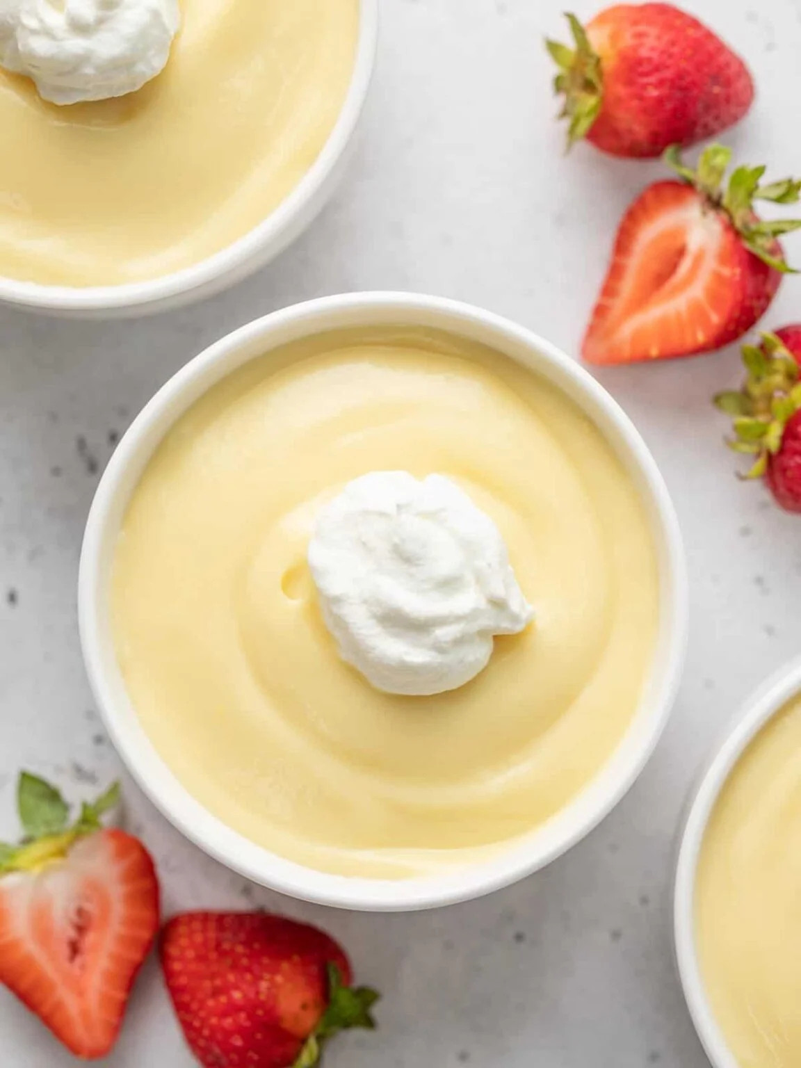 A white ceramic tub filled with yellow vanilla pudding and topped off with whipped cream. There are sliced strawberries nearby, and the edges of two other cups. 