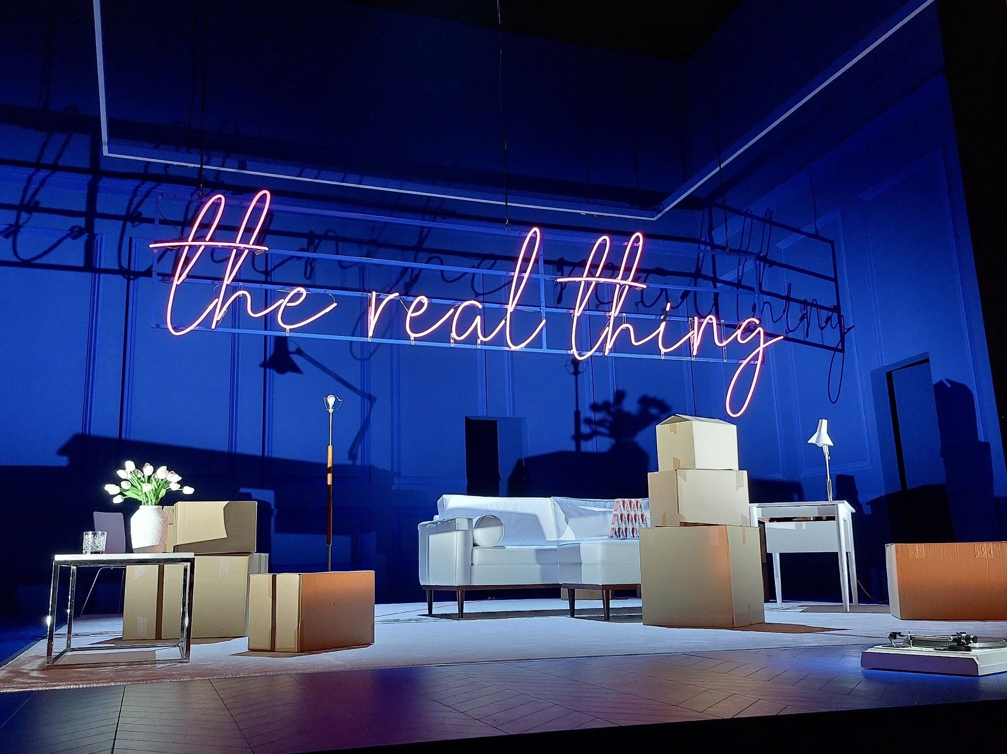 Set of The Real Thing. Scene of a minimalist living room with boxes still to be unpacked, blue stage lighting and the words The Real Thing about the set in pink neon lighting.