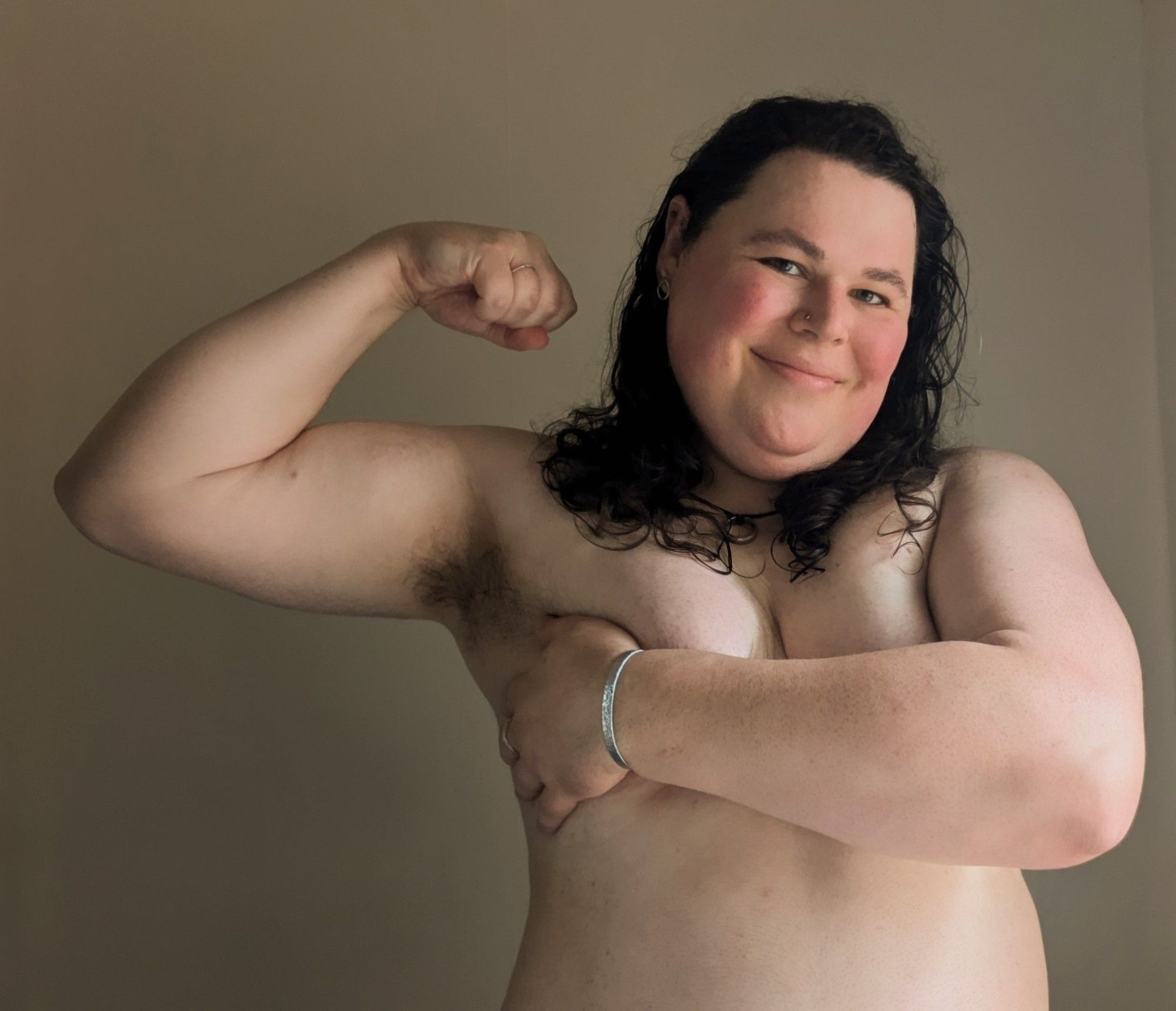 Alex, a large white trans woman, flexing with one arm and covering herself up with another. Her arms look big and strong and her smile is warm and soft.