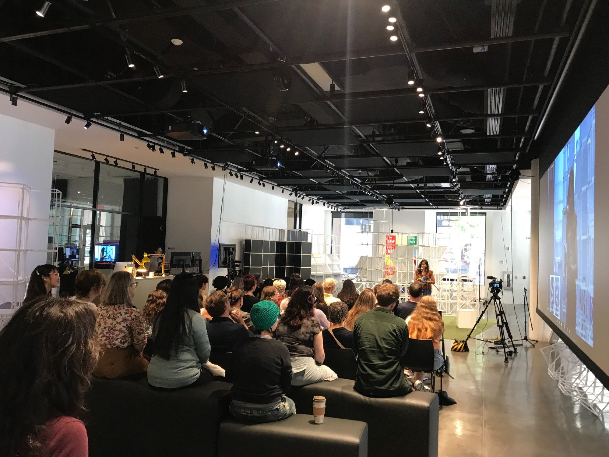 Photo of the Headlight relaunch at Concordia's 4th space: six rows of audience members with their backs to the camera and, at the background, Manahil Bandukwala performing at the mic, facing the audience.