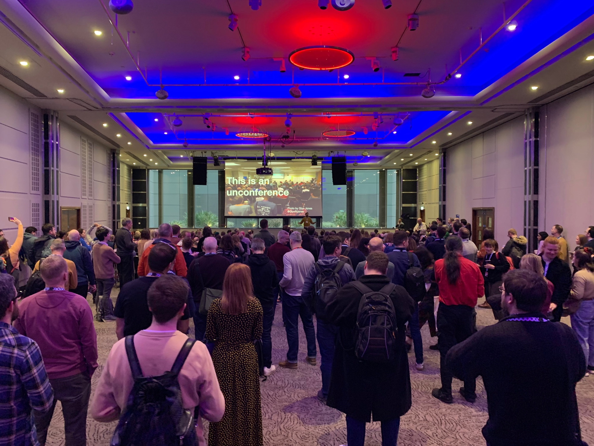 A large crowd facing the front of the large room used for UKGovCamp for the introduction to the day. There is a screen with a PowerPoint which says “This is an unconference”