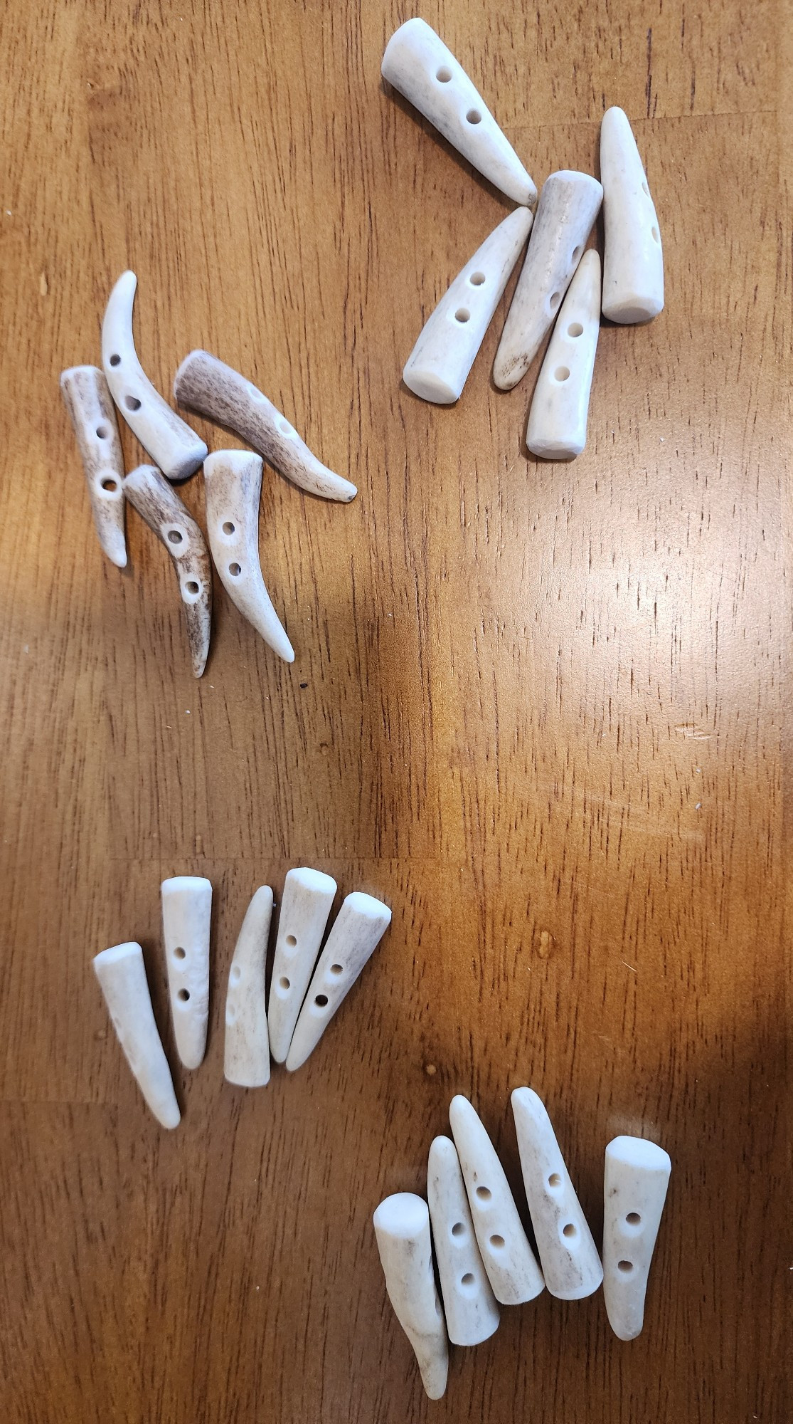 four sets of two-hole buttons made from deer antler tips are grouped on top of a wooden desk.  each set varies slightly in texture, color and size. some are a pale bone white and some are streaked with deep brown. some are very curved or crooked, showcasing the variability of the natural material.