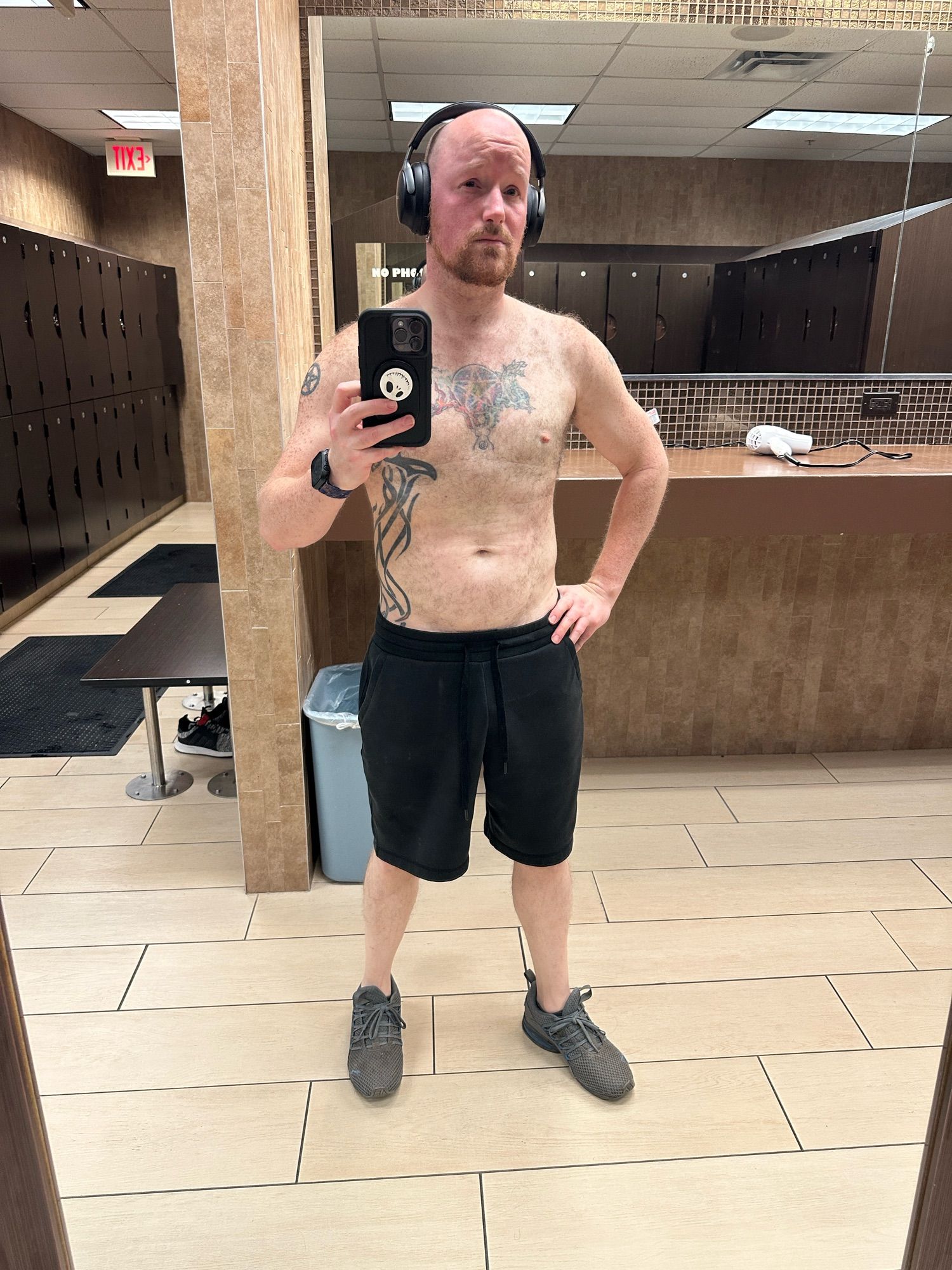 A selfie of me in my gym locker room.  I’m shirtless, wearing a pair of black shorts, and grey and blue sneakers, with a pair of headphones on.