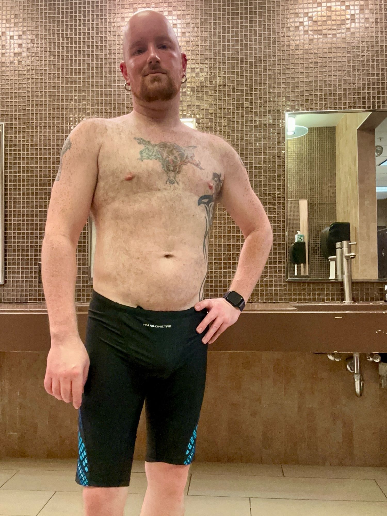 A selfie of me in the gym locker room.  I am wearing a pair of black swim jammers with pale blue diamond patterns going down the legs of the pant near the knee.