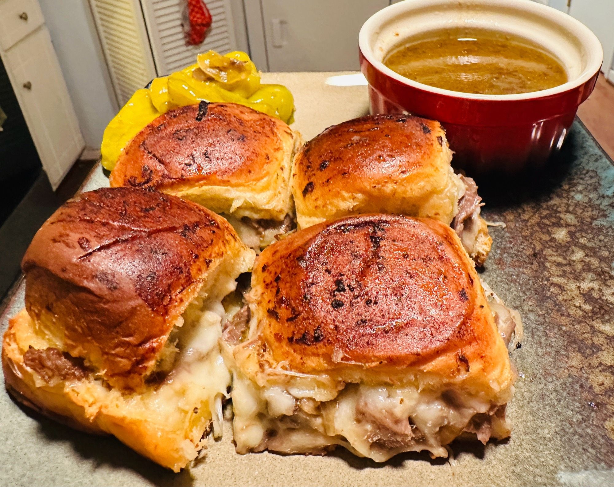 A picture of four sliders on a stoneware plate.  With a cup of left over jus( roast gravy/drippings) for dipping.  The cheese is just oozing out of the sliders 🤤