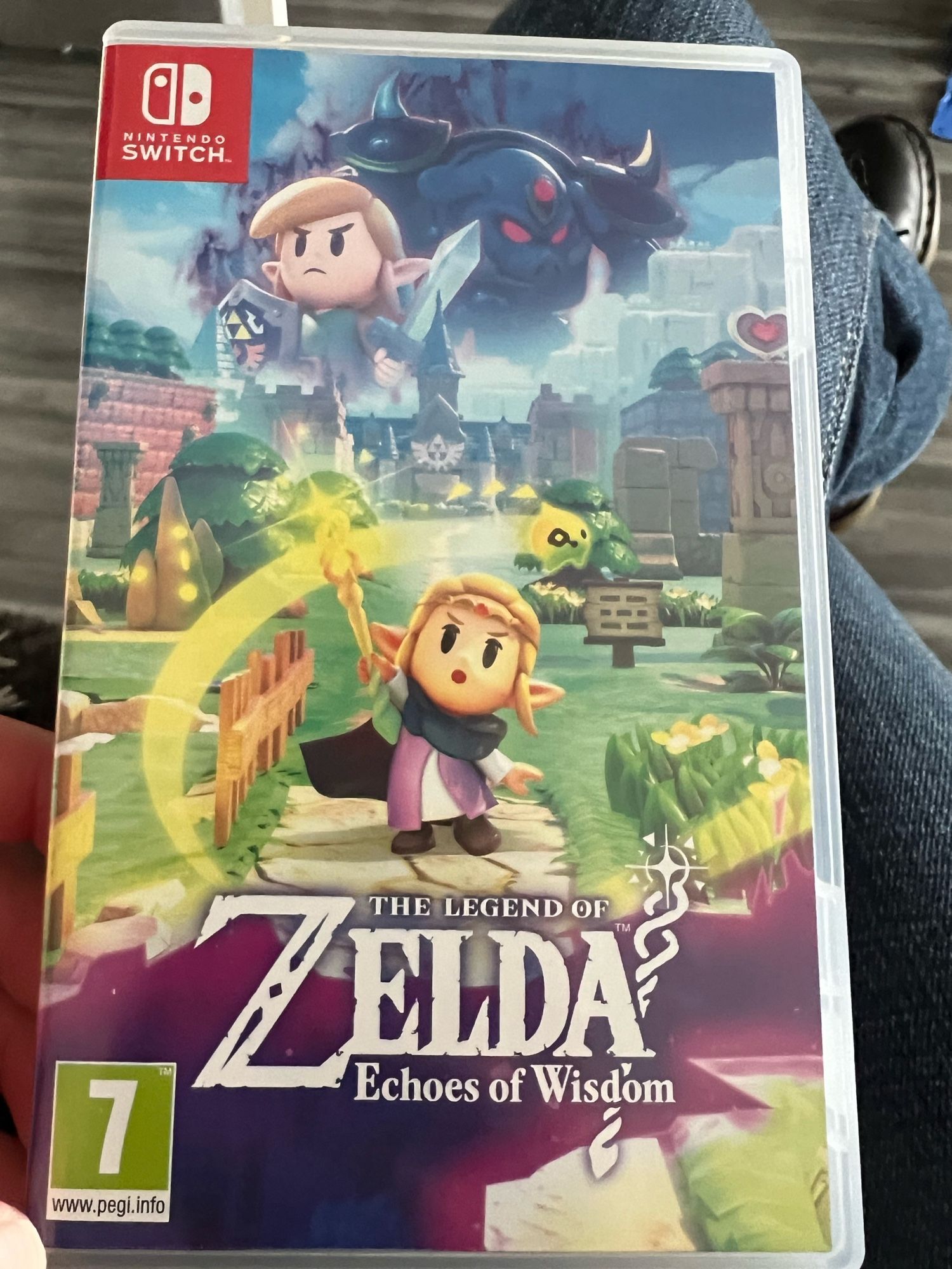 The Legend of Aelda Echoes of Wosdom game case. It has an image of Princess Zelda  holding up a staff of some sort, with an image of Hyrule behind her. Above her is a faded image of Link and I assume Ganondorf. The design is the same cartoony design as the Link’s Awakening game.