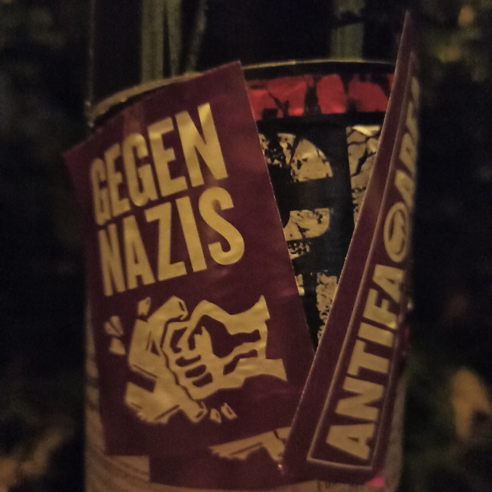 Ein Foto. Im Dunkeln aufgenommen 
Zu sehen sind zwei Antifa-Sticker die einen Fascho-Sticker überklebt haben 
Der eine Sticker zeigt eine Faust die ein Hackenkreuz zerschlägt darauf zu lesen ist Gegen Nazis der andere Sticker zeigt die Aufschrift Antifa Area