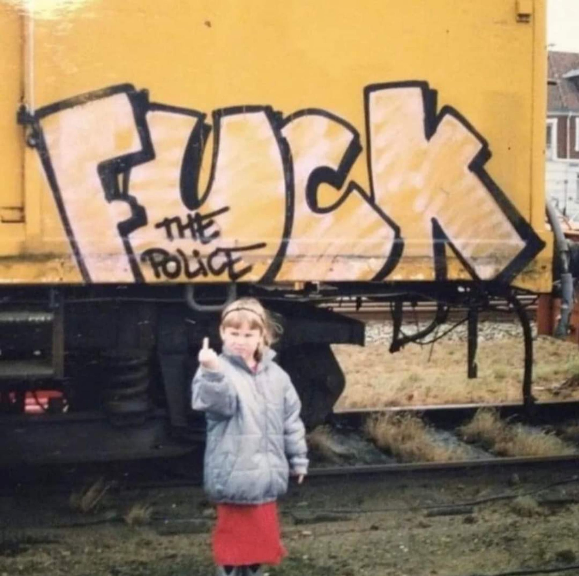 Foto. Zeigt ein Mädchen das den Stinkefinger zeigt. Im Hintergrund ist eine Lokomotive zu sehen. Darauf ein Graffiti. Zu lesen ist Fuck the Police