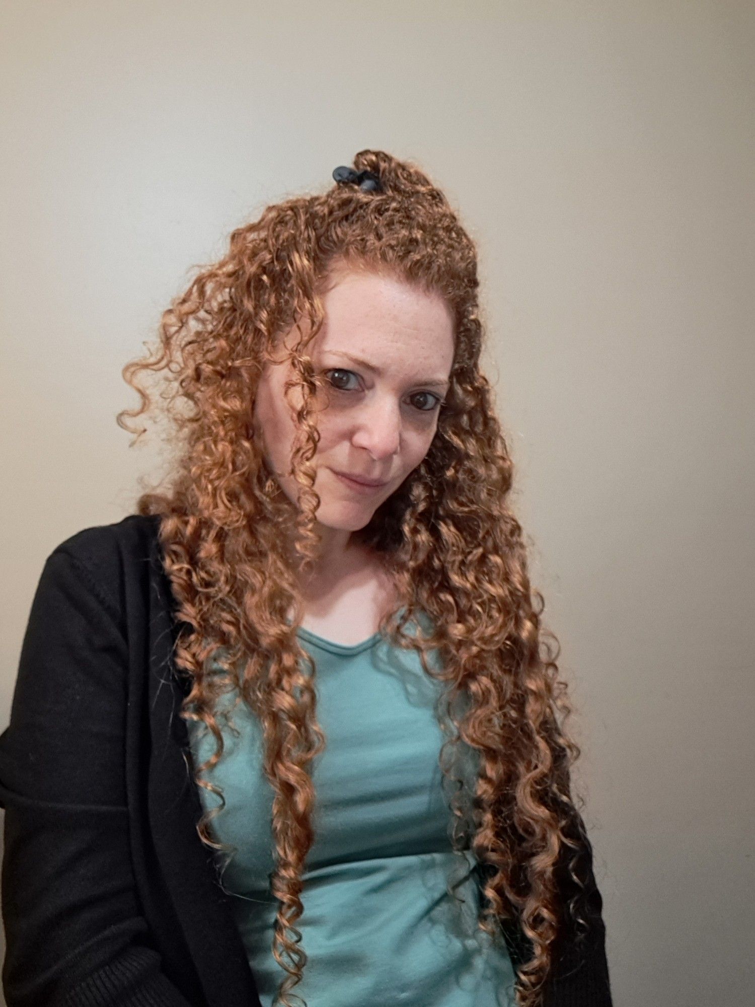 Some of her spiral curly hair is held to the top of her head with a clip, the rest piles over her shoulders.  She's wearing a teal shirt and black cardigan