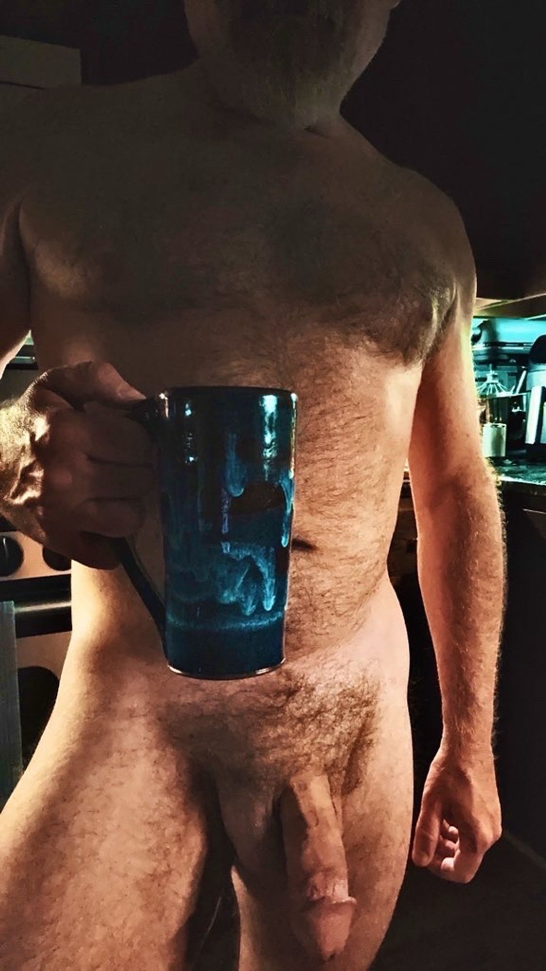 Furry naked Dad standing in the kitchen holding a blue ceramic mug while his soft thick cock hangs between his legs.