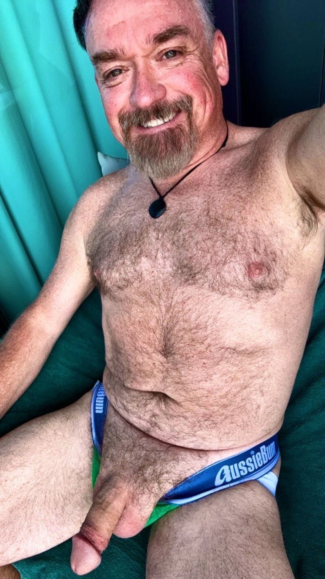 Furry Dad sitting in a chair with his thick floppy cock sticking out of his green Aussiebum jockstrap.