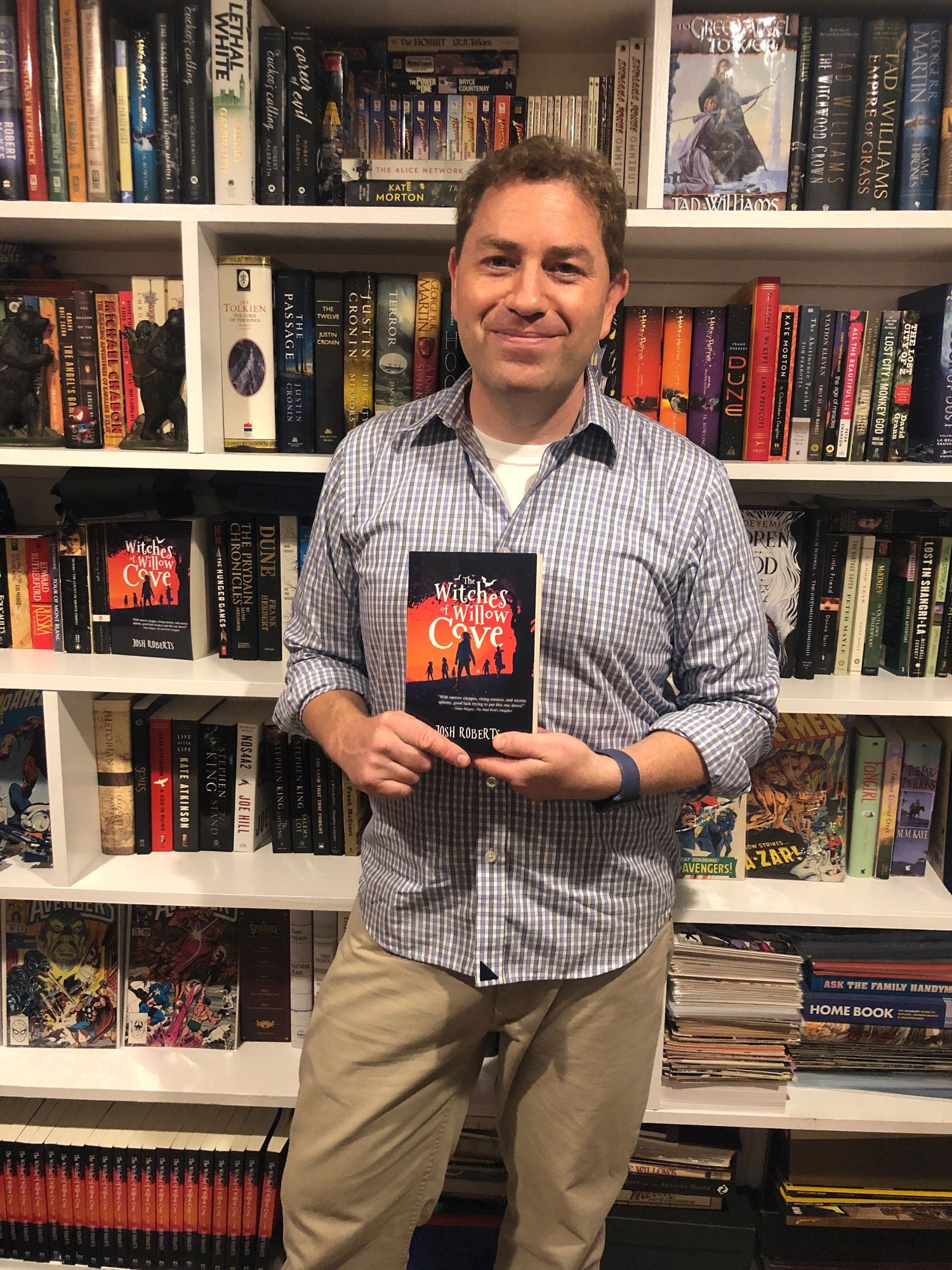 Josh Roberts standing in front of a book shelf holding a copy of The Witches of Willow Cove.