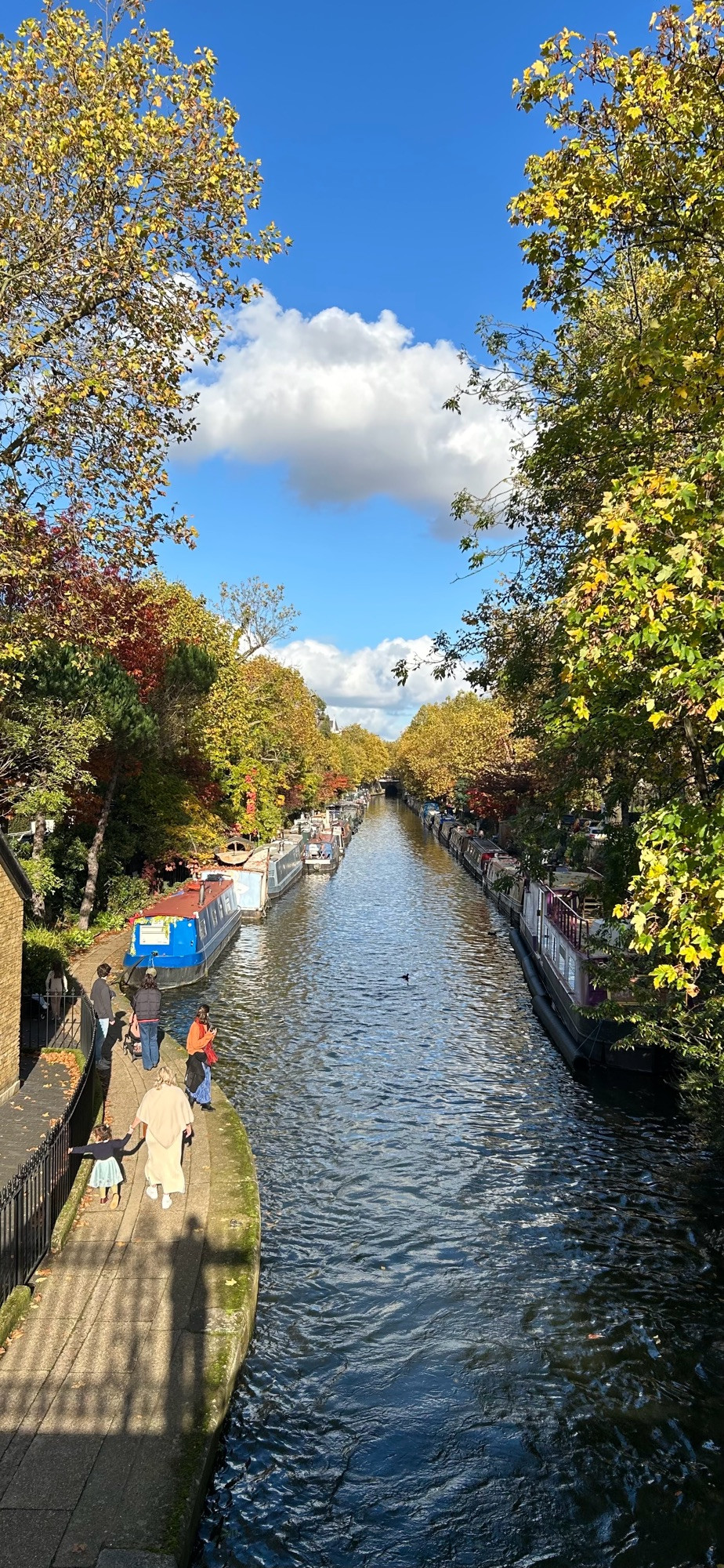 Photo of a canal in autumn. The canal is wide, with narrowboats (barges) moored on either side. The banks of the canal are lined with trees, whose leaves are a variety of shades of green, gold and red. On the towpath to the left, there are a few pedestrians enjoying the scene in the sunshine, and we can see the shadow of the photographer. In the distance, we see the portal of a tunnel through which the canal passes. The sky is bright blue, with some puffy clouds.