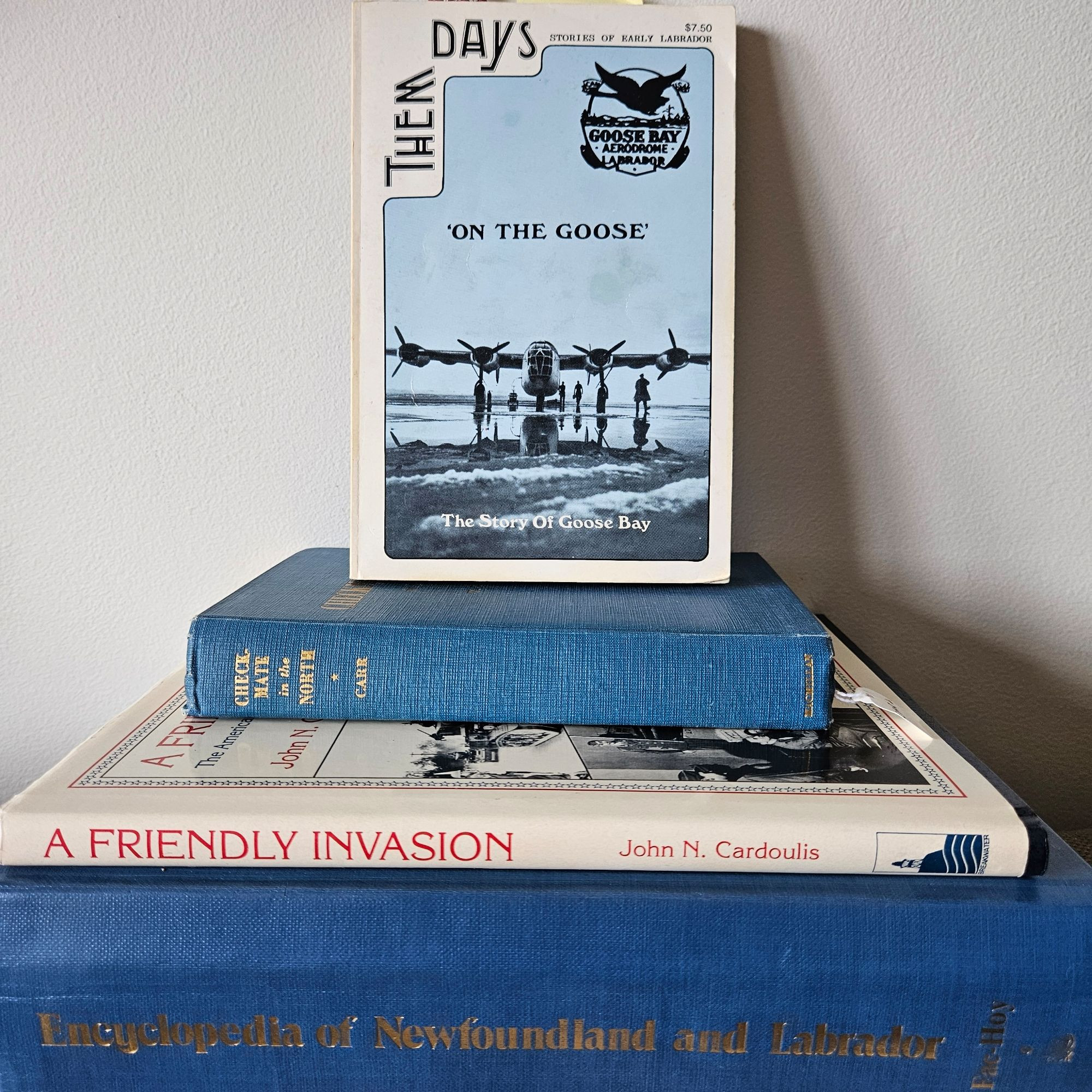 A stack of books against a white background. On top, with the cover facing forward is Them Days "On the Goose" the story of Goose Bay with a picture of a B-24 Liberator aircraft. Under that, stacked with their spines facing forward are: Checkmate in the North by W.G. Carr, A Friendly Invasion by John N. Cardoulis, and Encyclopedia of Newfoundland and Labrador, Fac-Hoy.