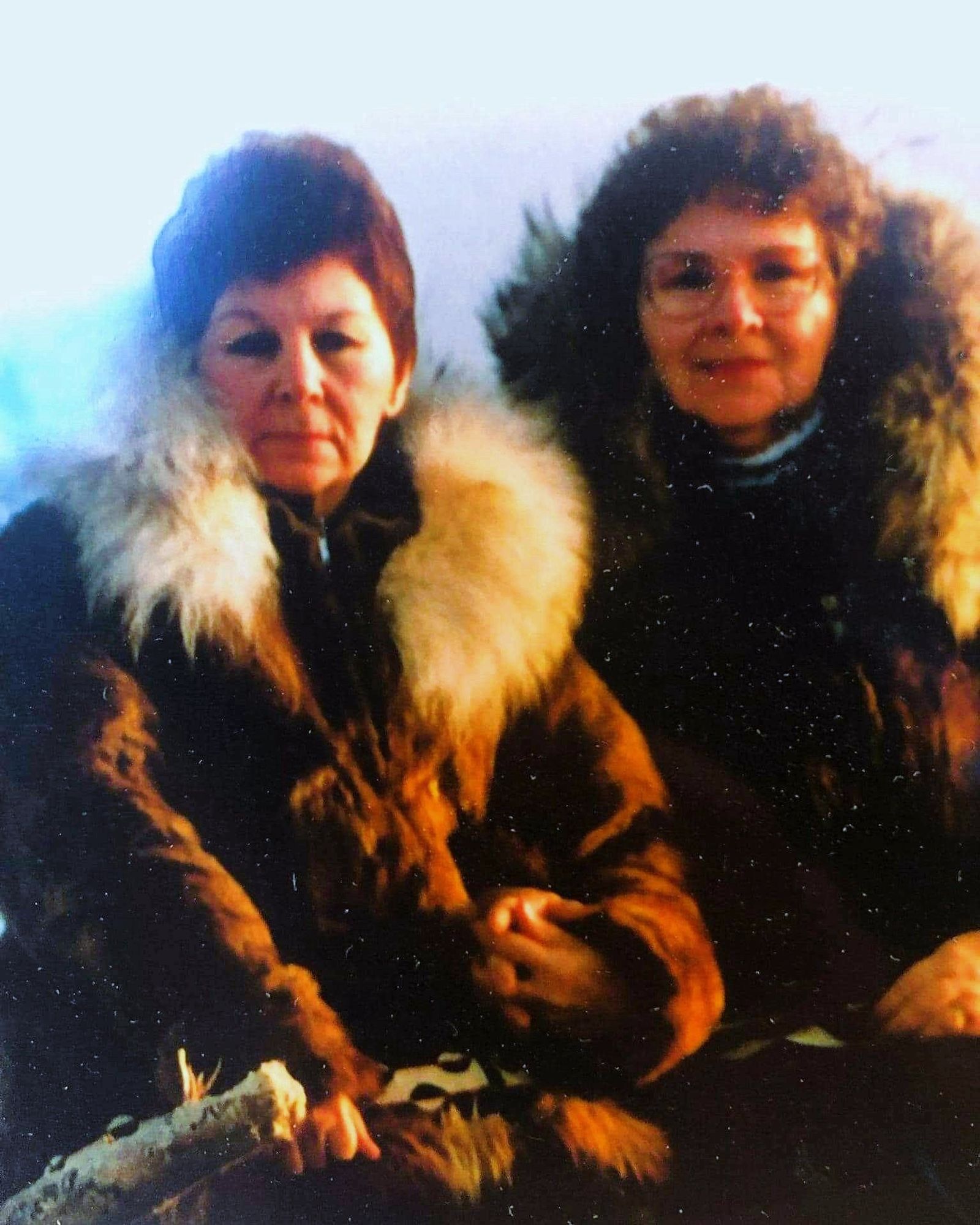 An old photo of 2 middle-aged Inuuk women in brown fur parkas, holding hands. The woman on the left with the lighter collar on her parka is Elsa. On the right, wearing glasses, is her sister Anna.