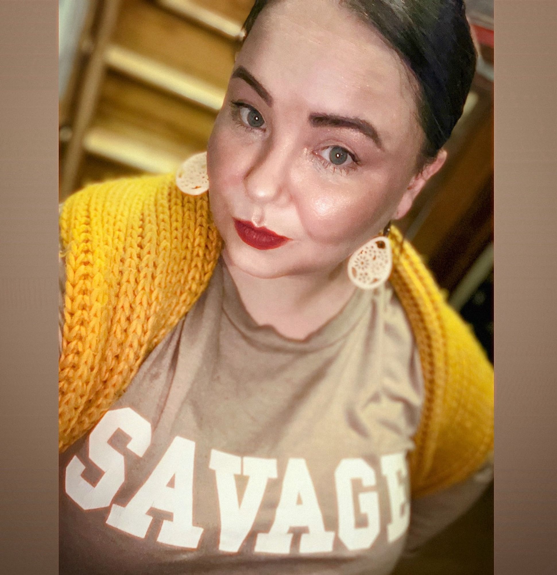 A selfie of C with a serious expression. She is wearing ivory earrings carved by an Inuk artist & a tan shirt with the word SAVAGE in block letters prominently across the chest. She has a "Square Yellow" chunky knit scarf draperd across her shoulders (sort of a golden mustard shade)