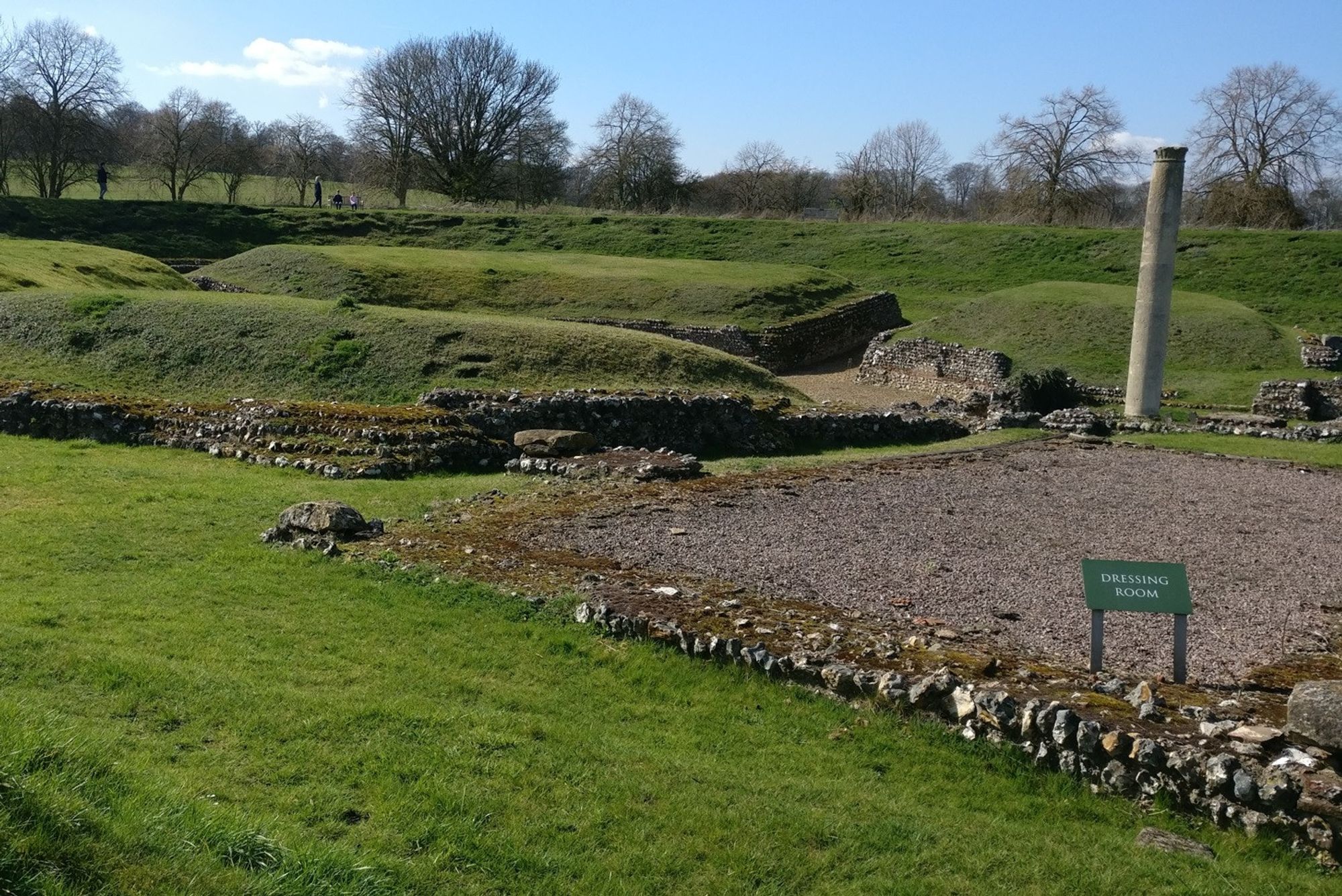 The site of some historical ruins, with grass banks in the background. In front some recognisably building shaped remains (or at least their foundations) with a sign saying "Dressing Room". A lone column stands forlornly at the rear.