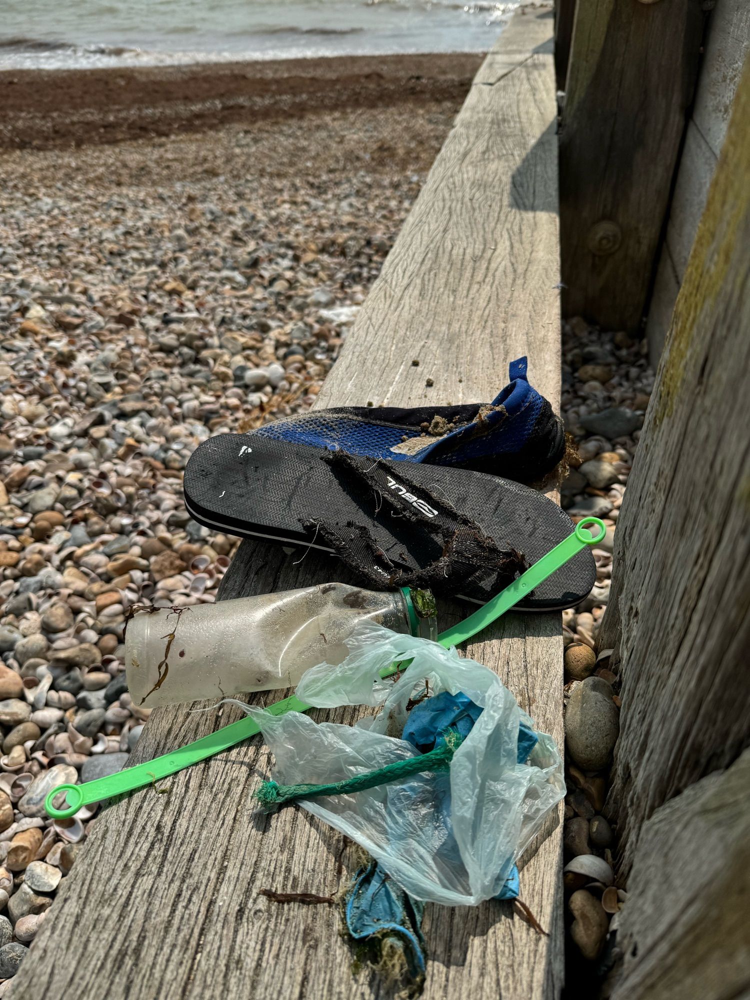 Beach shoes plus plastic items including bottles, bags, string and ties found on a pebble beach during a beach clean.