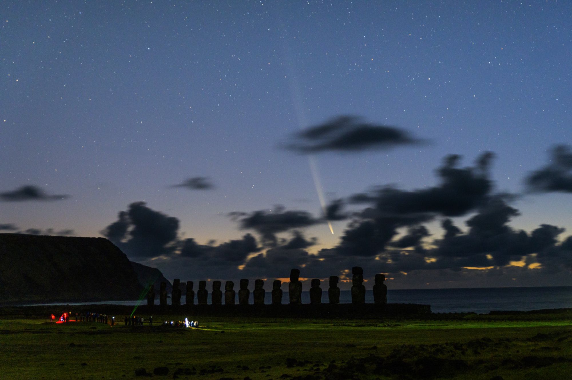 A comet with a long tail in twilight skies with some clouds and Easter Island statues in the foreground.