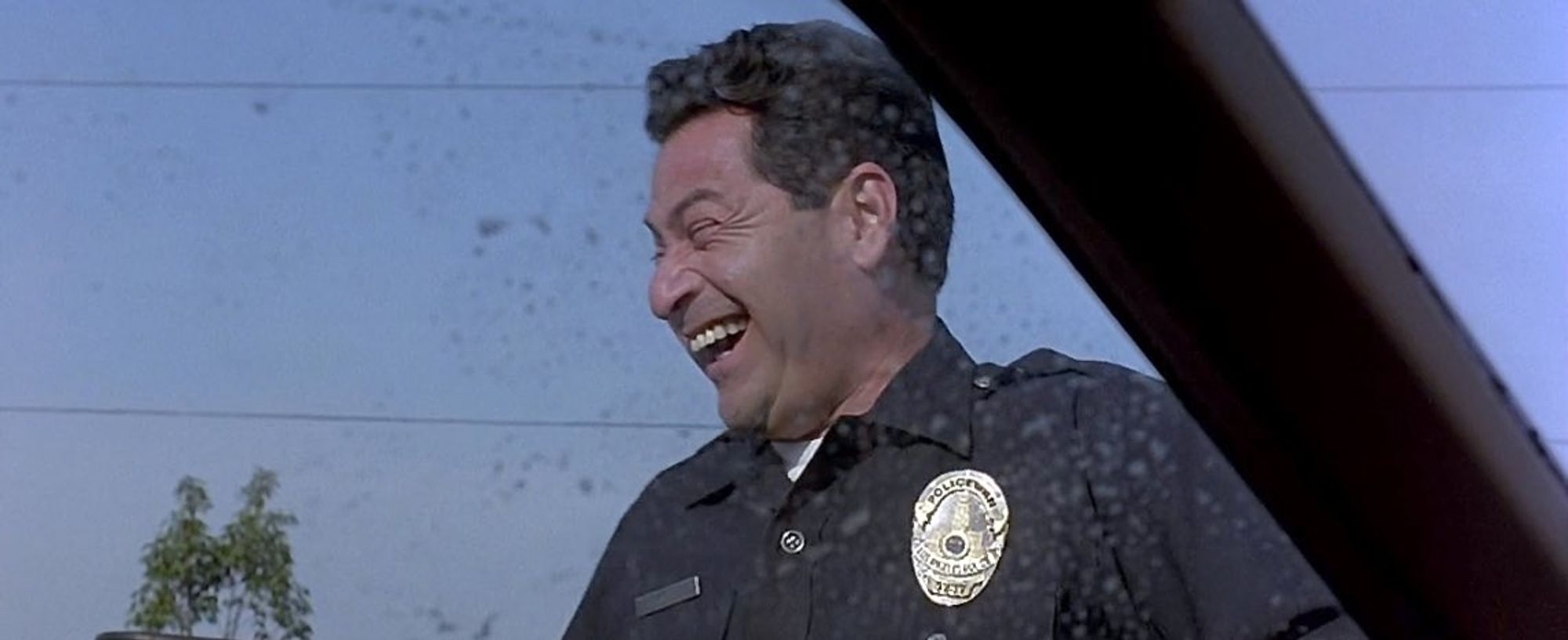 A still image from the movie. The Big Lebowski showing a cop laughing. In the scene he is mocking the main character for thinking the police would investigate his stolen car.