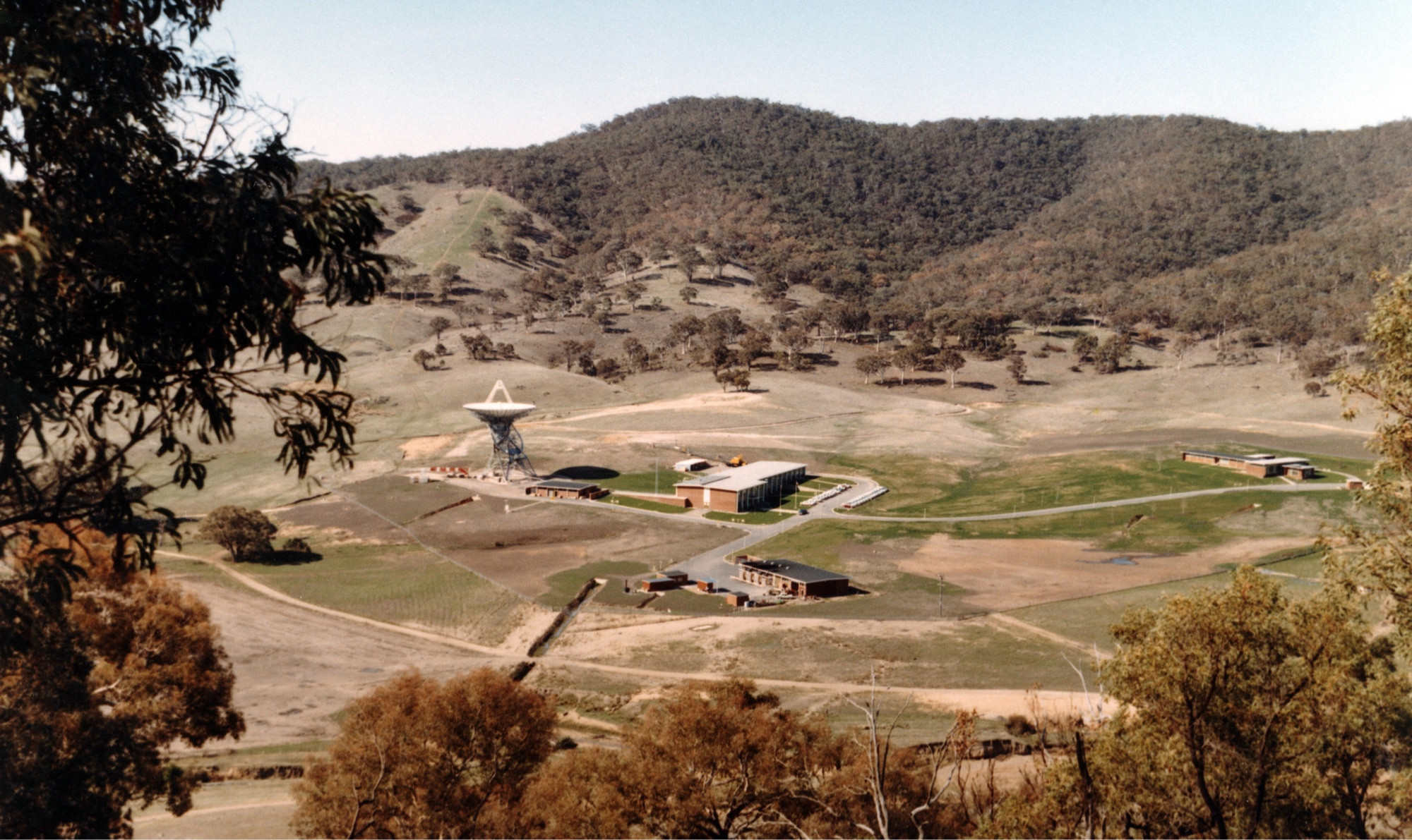 Elevated view of the Tidbinbilla Deep Space Instrumentation Facility 42 in 1965. A single antenna dish is near some buildings set in an open green valley surrounded by hills covered in trees.