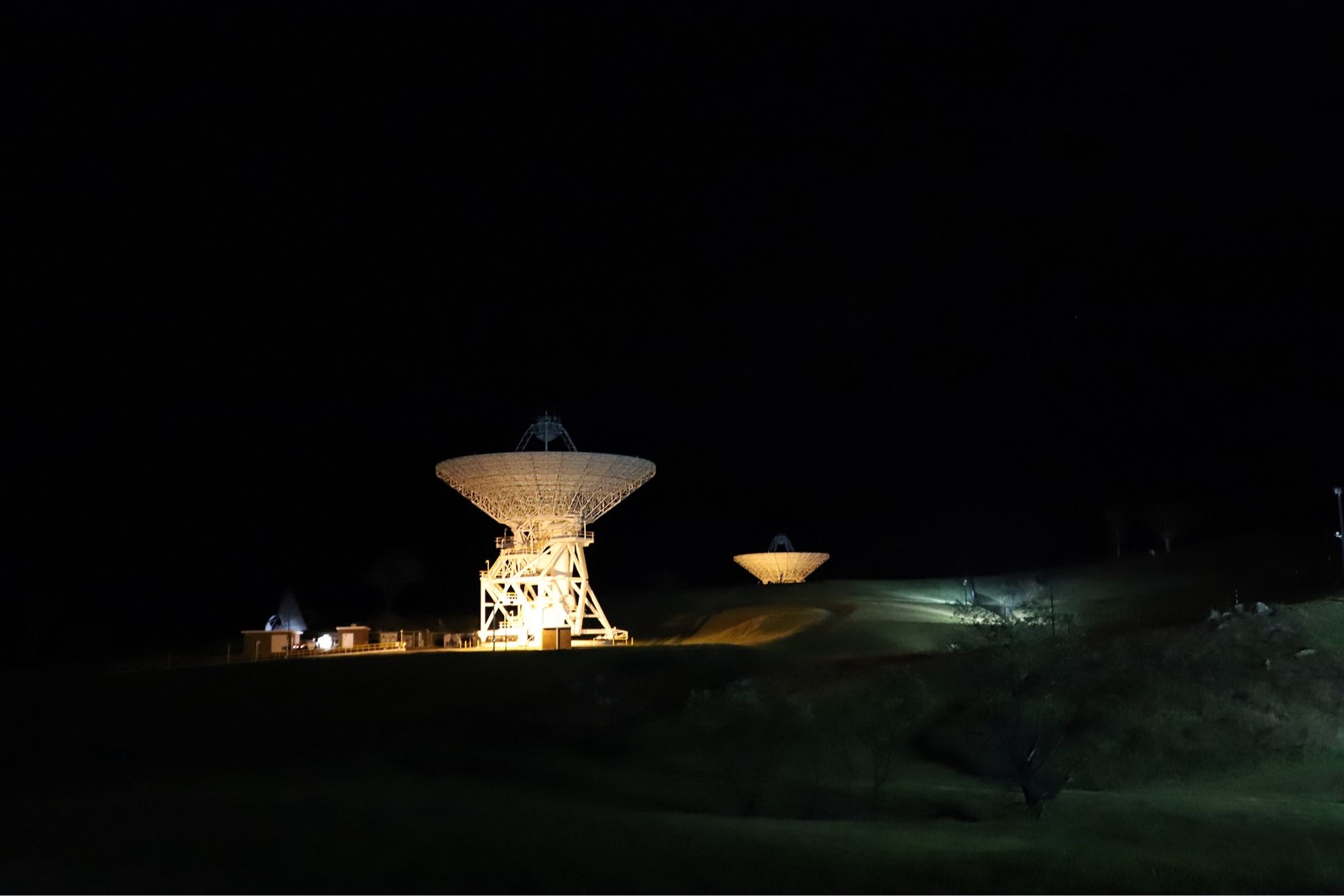 Two deep space antenna dishes are lit by foreground lights, and are set against a black night sky. Deep Space Stations 34 and 35 are located at the Canberra Deep Space Communication Complex, Australia.