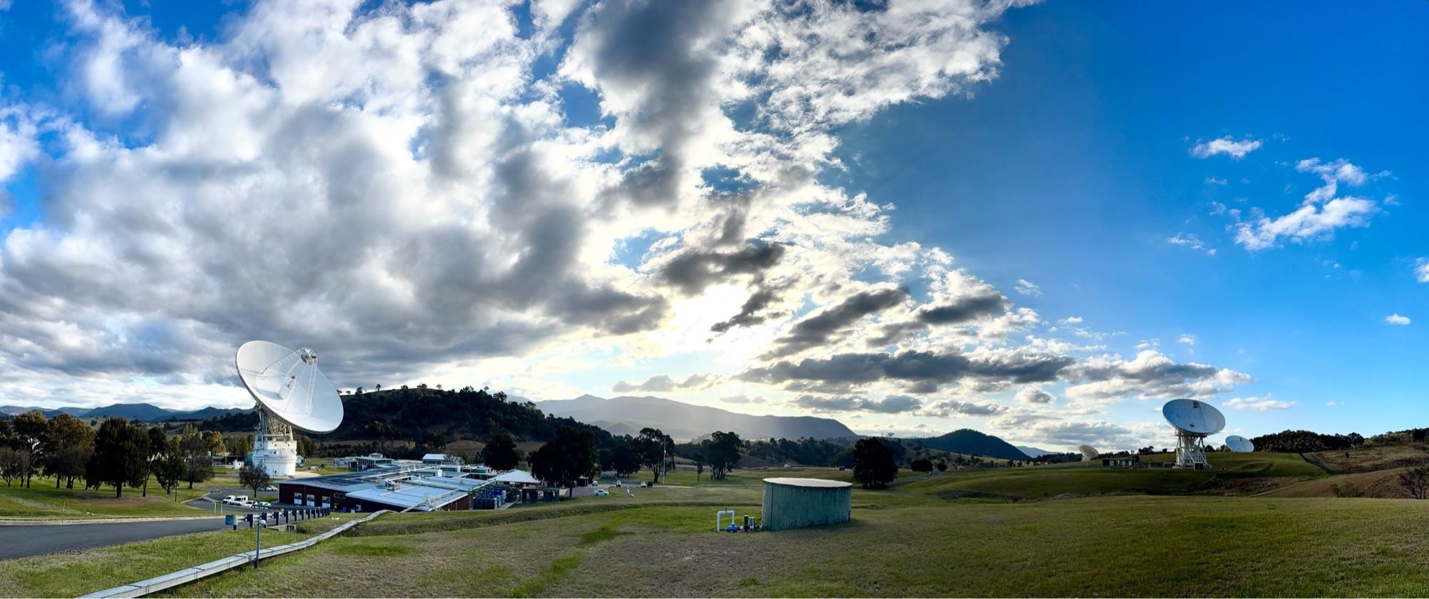 Panoramic view of deep space antenna dishes pointing skyward. Hills and setting sun in a cloudy blue sky.
Deep Space Station 43 on left in contact with NASA’s Voyager 1 spacecraft, and on the far right, antenna dishes Deep Space Stations 34 and 35 linked as an array to receive data from Voyager 2.