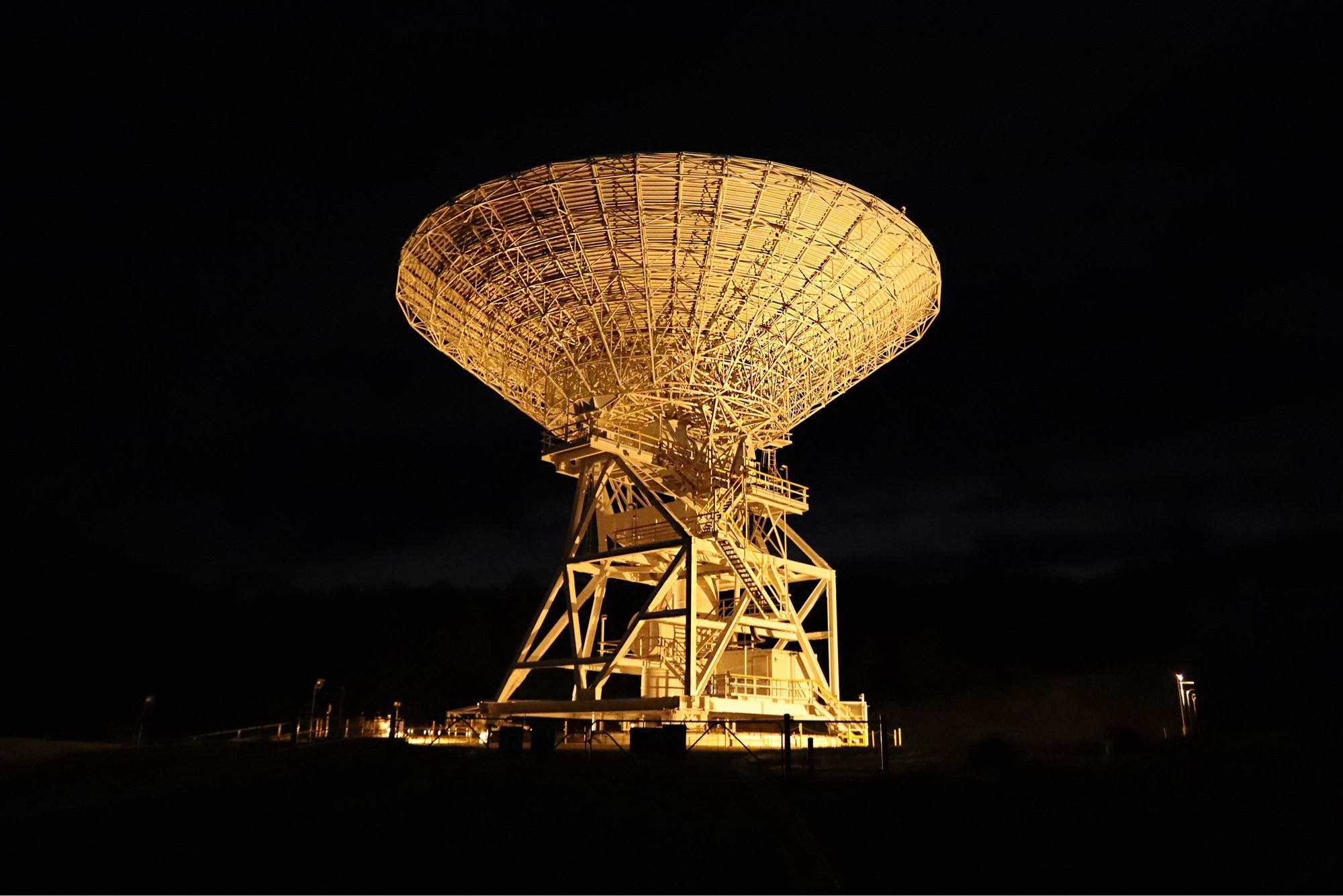 Bathed in orange light, a deep space antenna dish stands against the black night sky. Deep Space Station 34 is located at the Canberra Deep Space Communication Complex, Australia.