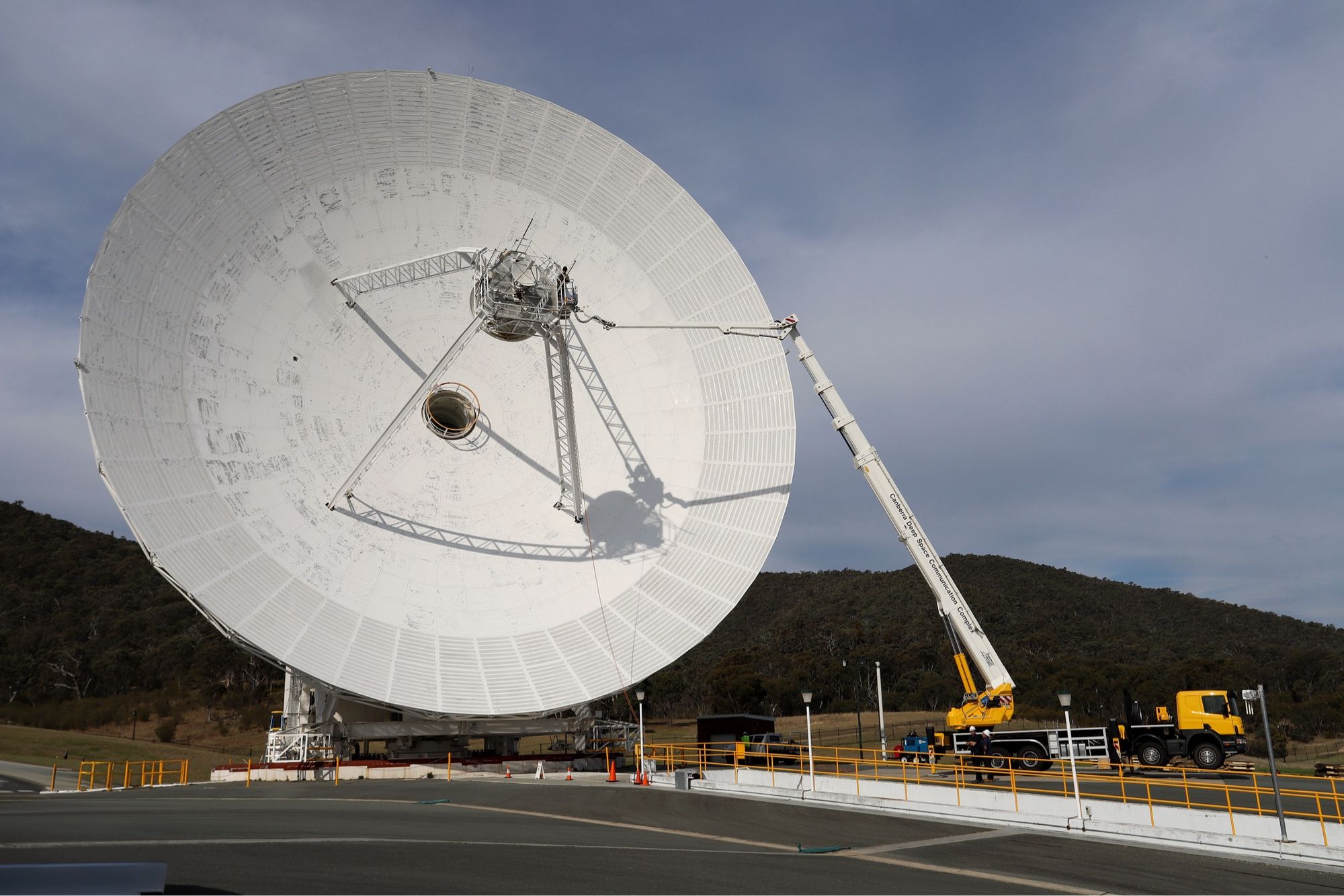 A truck mounted elevated work platform is extended towards the centre section of a 34-metre diameter antenna dish which has been tilted over low to the ground. Deep Space Station 34 at the Canberra Deep Space Communication Complex, Australia. Part of NASA's Deep Space Network.