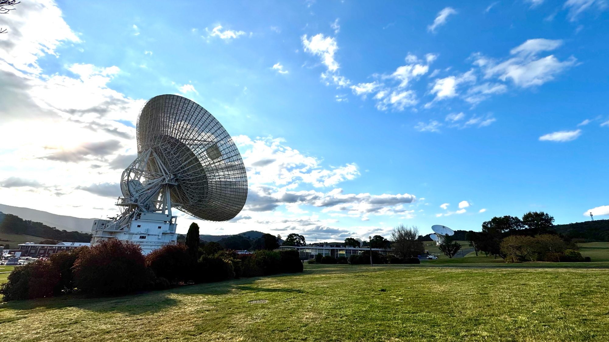 Panoramic view of deep space antenna dishes pointing skyward. Hills and setting sun in a cloudy blue sky.
Deep Space Station 43 on left in contact with NASA’s Voyager 1 spacecraft, and on the far right, antenna dishes Deep Space Stations 34 and 35 linked as an array to receive data from Voyager 2.