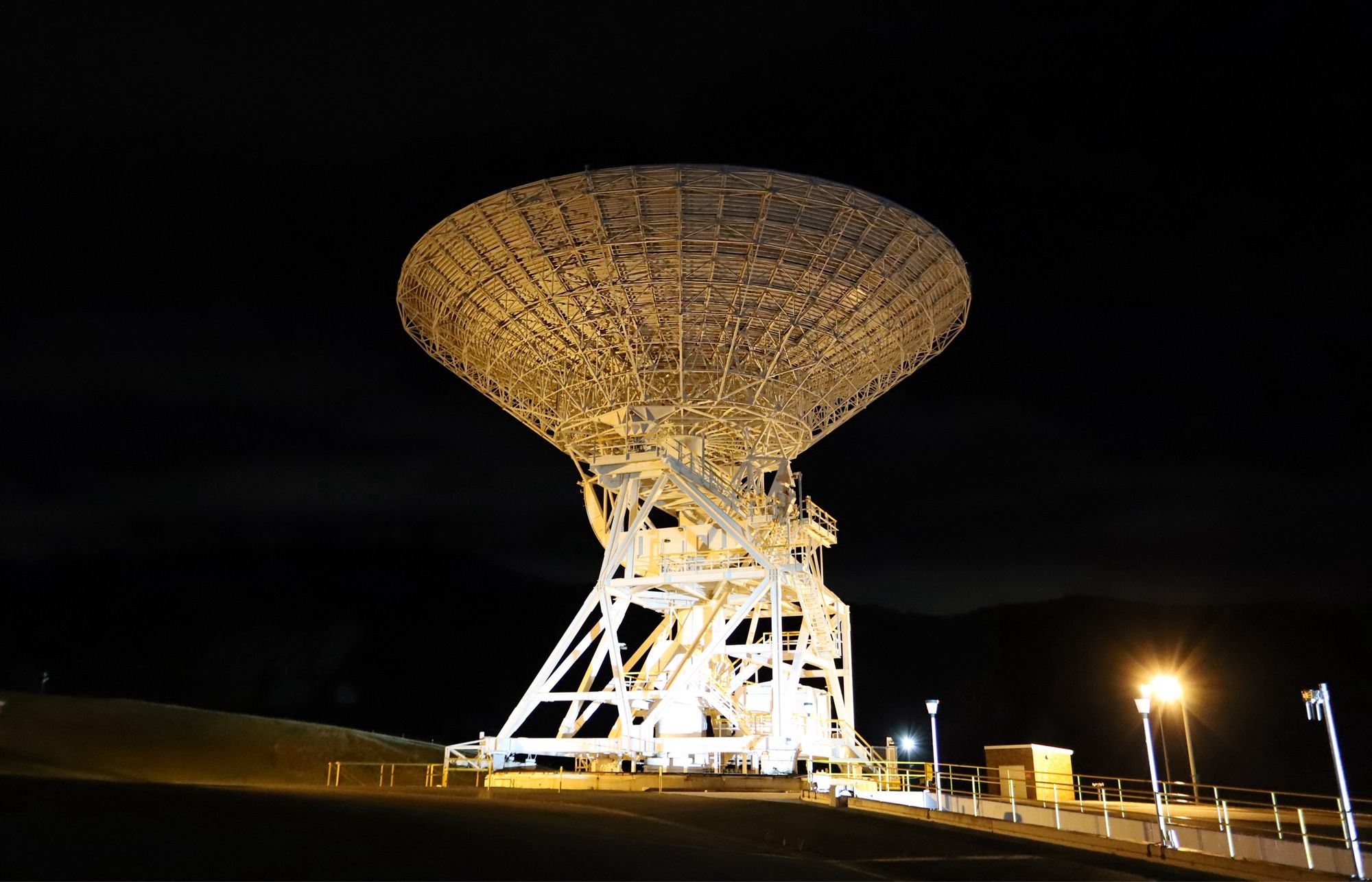 Bathed in orange and white light, a deep space antenna dish stands against the black night sky. Deep Space Station 34 is located at the Canberra Deep Space Communication Complex, Australia.