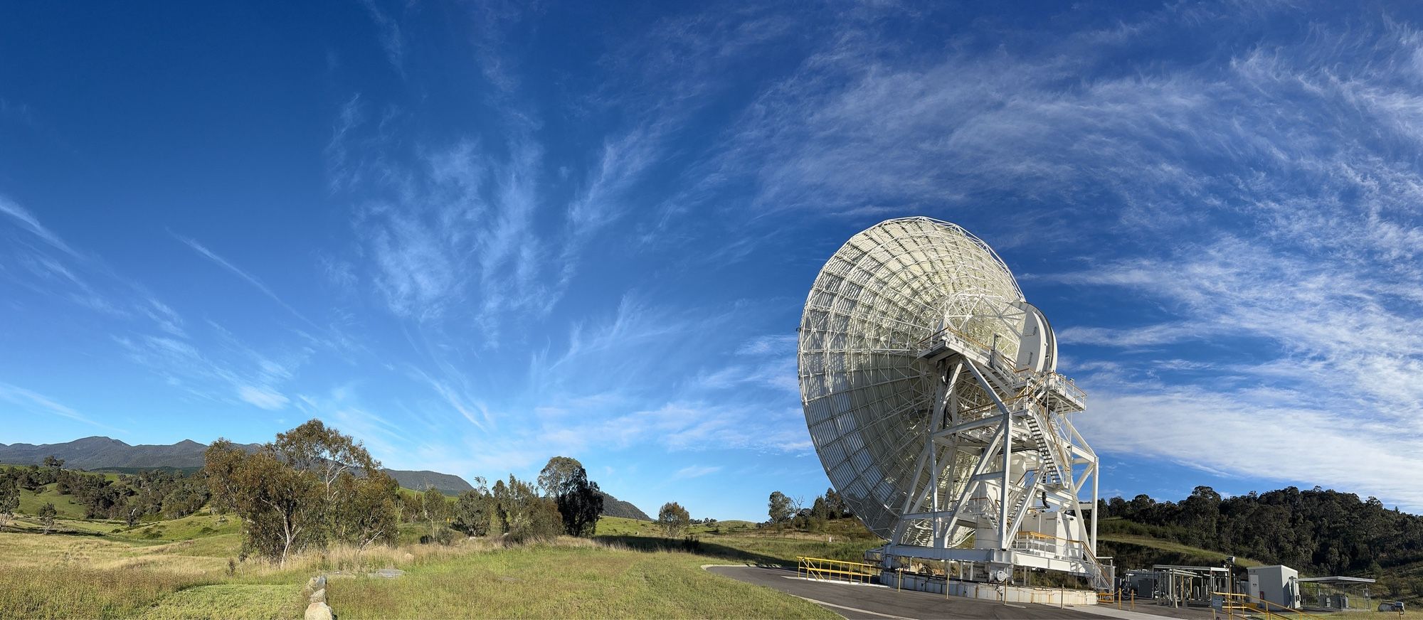 Wispy clouds against a blue sky above bushland. The foreground is dominated by a large deep space antenna dish, pointing to the horizon. Antenna Deep Space Station 36 at the Canberra Deep Space Communication Complex, Australia.