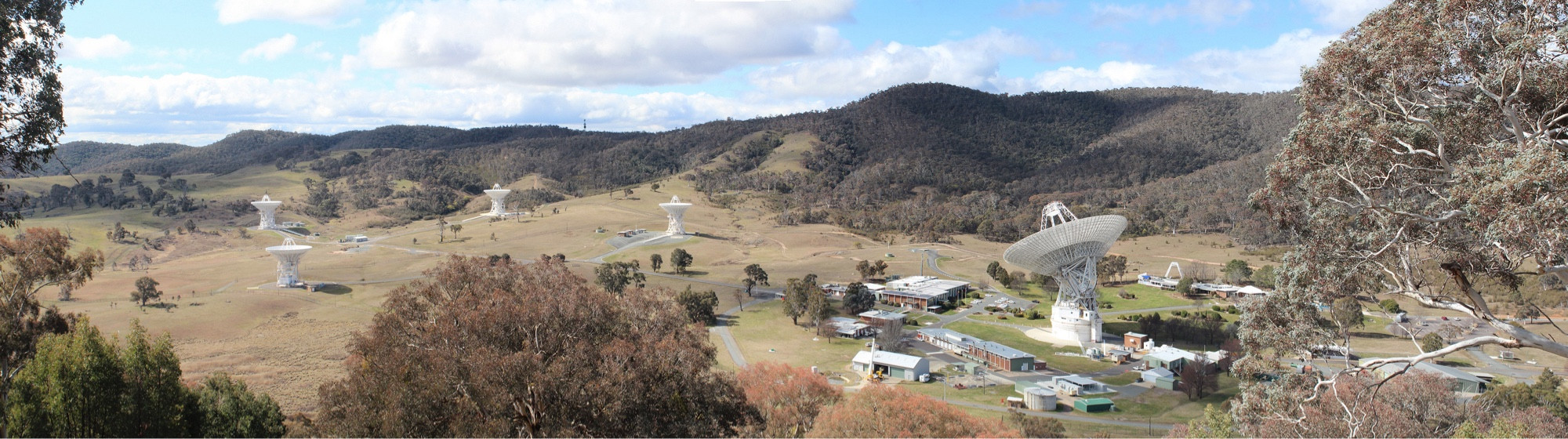 Elevated view of the Canberra Deep Space Communication Complex as it stands today. Multiple antenna dishes and buildings set in an open green valley surrounded by hills covered in trees.