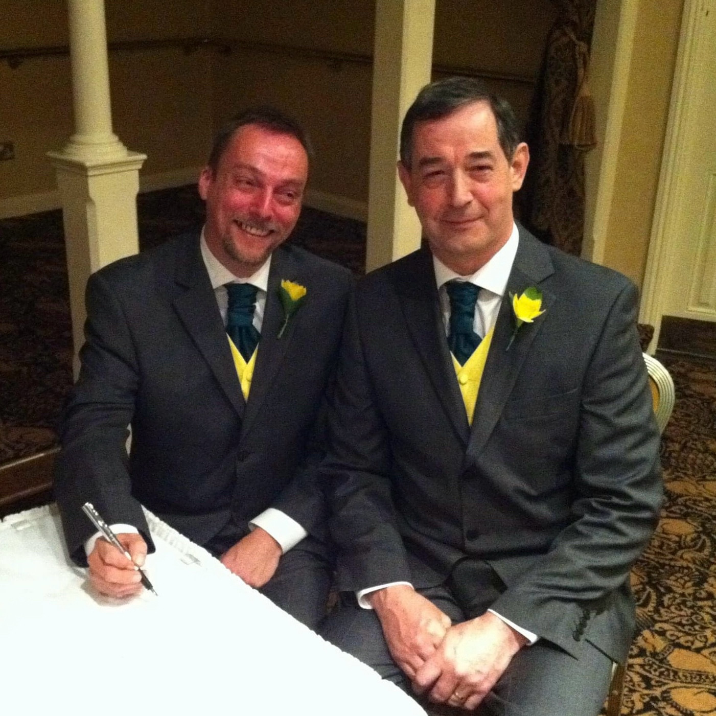 Two seated Caucasian men smiling in suits on their civil partnership day. Grey suits accented with green for shamrock of Ireland and Yellow for the daffodil of Wales.