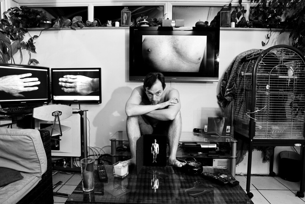 A naked man with Poland’s Syndrome in a room with a budgie. There are photos highlighting different aspects of his anatomy and how the syndrome alters the symmetry of the human form.