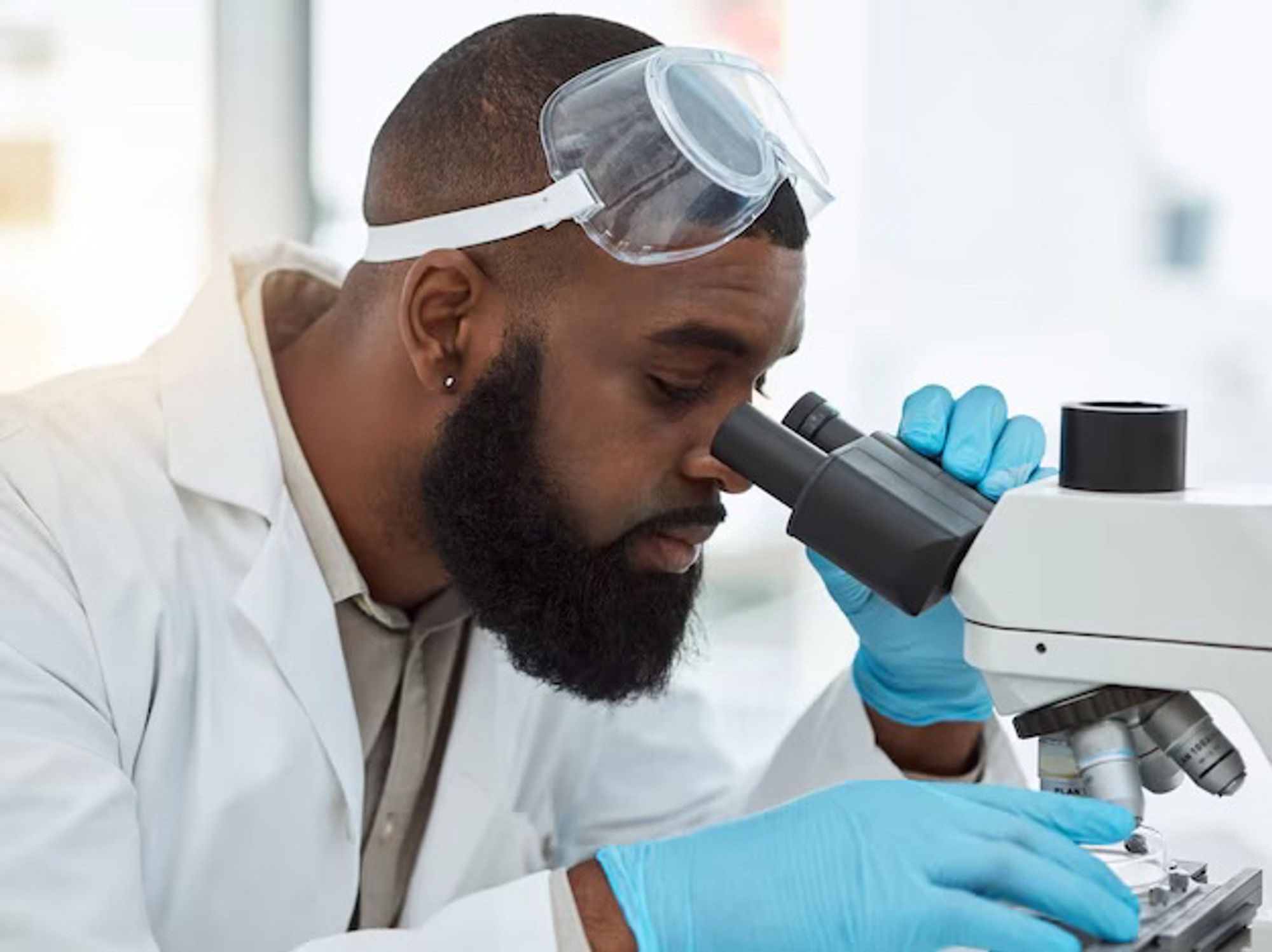 Photographie d’un scientifique noir en blouse, il porte des lunettes de sûreté, des gants et une barbe. Il regarde dans un microscope.